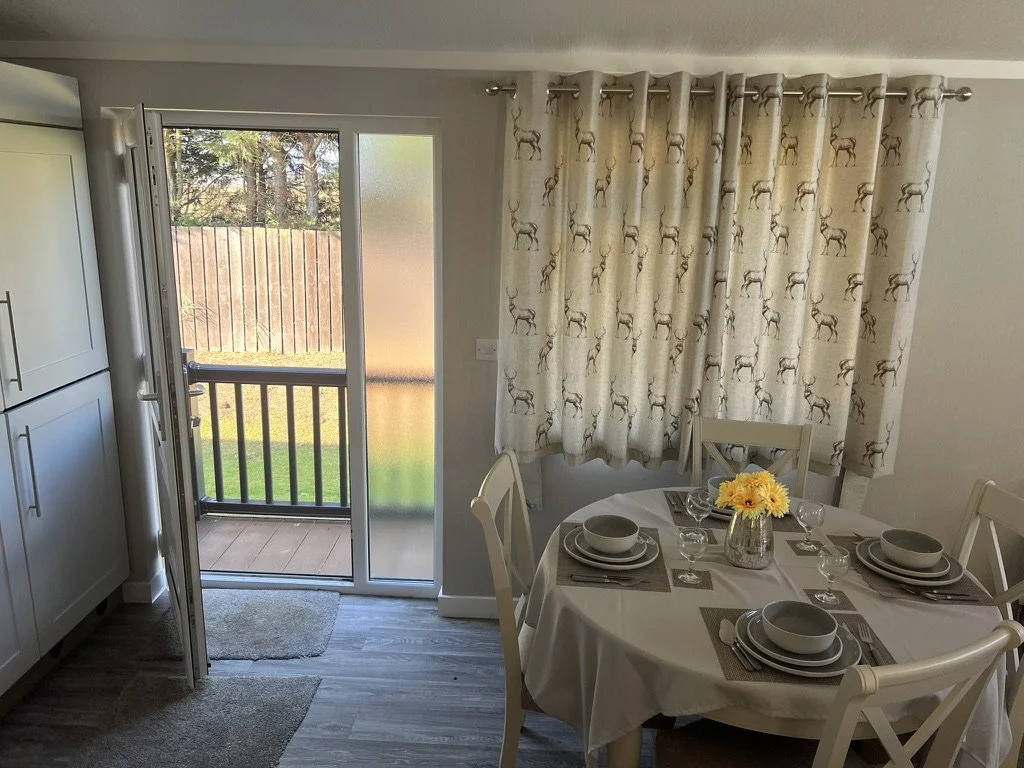 Dining area with a round table set with dishes, cups, and a vase of yellow flowers, next to a sliding glass door leading to a balcony and a window with curtains decorated with reindeer patterns.