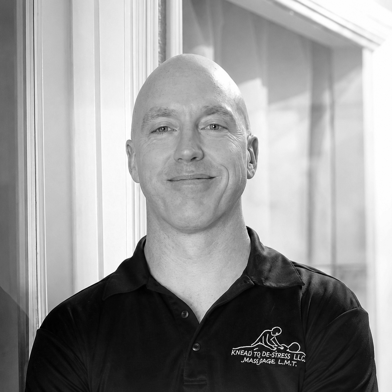 A smiling man in a black polo shirt with a logo and text about massage therapy standing in front of a window with natural light.