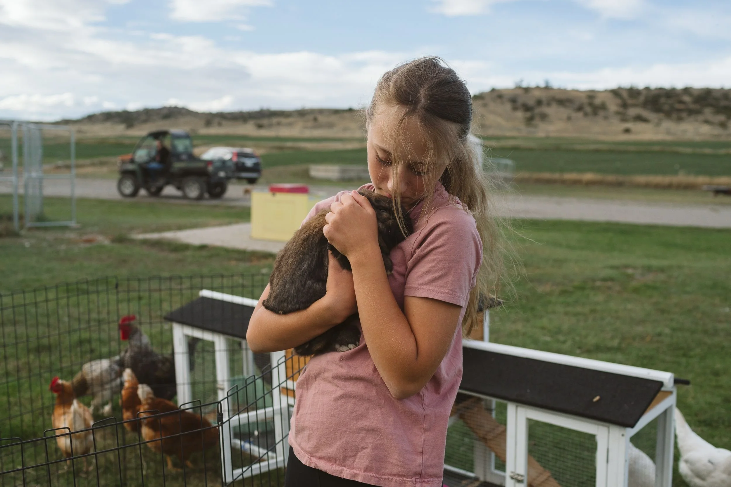 Young girl nuzzling a rabbit at a Montana ranch with rolling hills in the background, editorial ranch photography by Tori Pintar