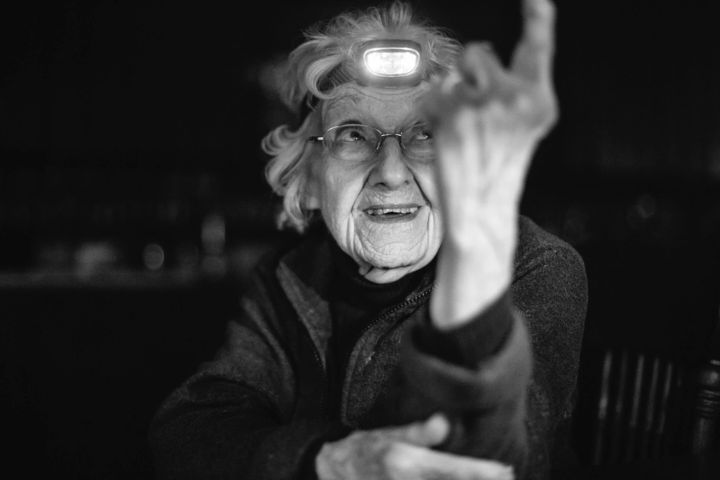 Black and white portrait of an elderly woman wearing a headlamp and gesturing expressively in the dark, documentary photography by Tori Pintar