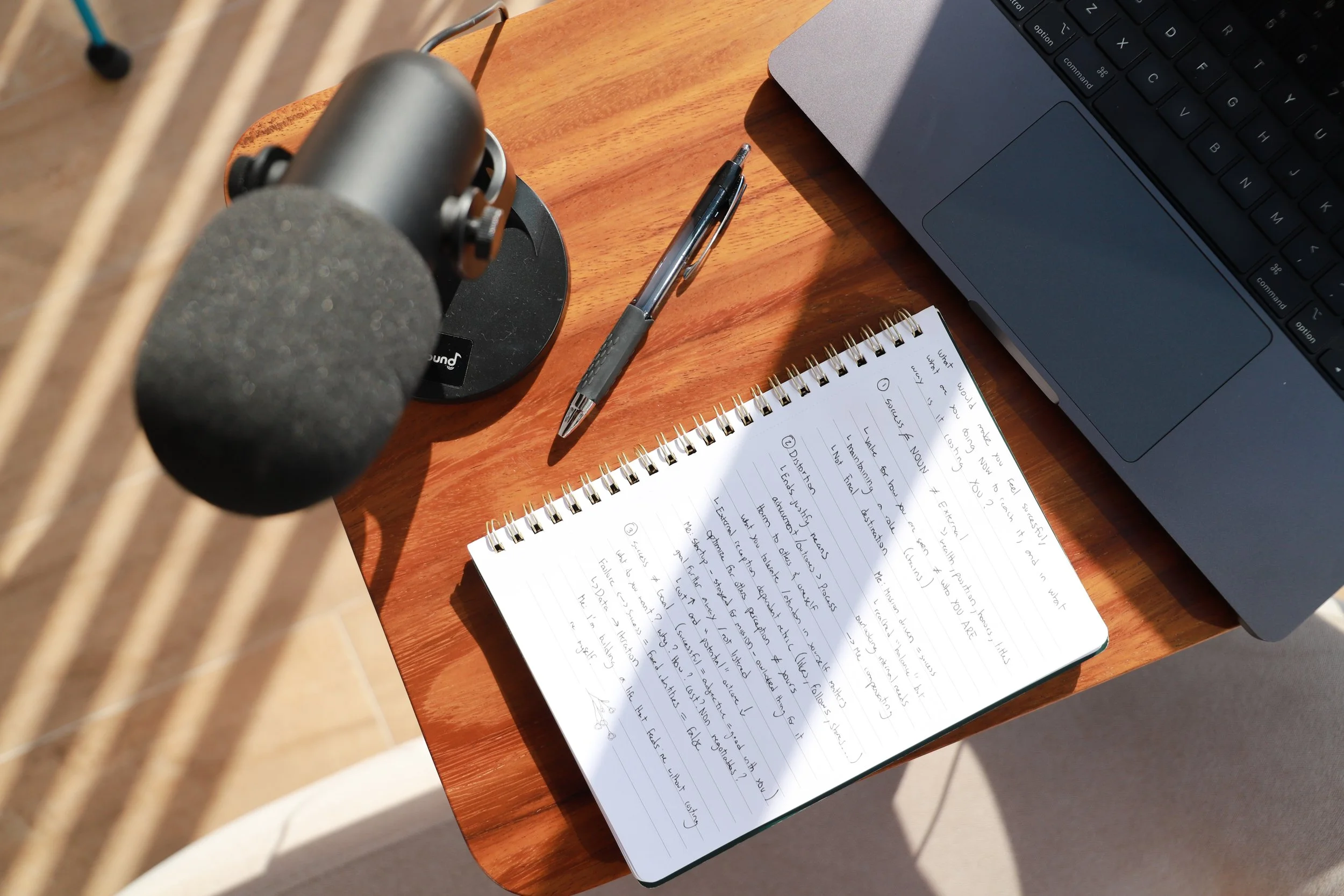 Podcast recording setup with a microphone, notebook, pen, and laptop in warm natural light, capturing a reflective creative process.