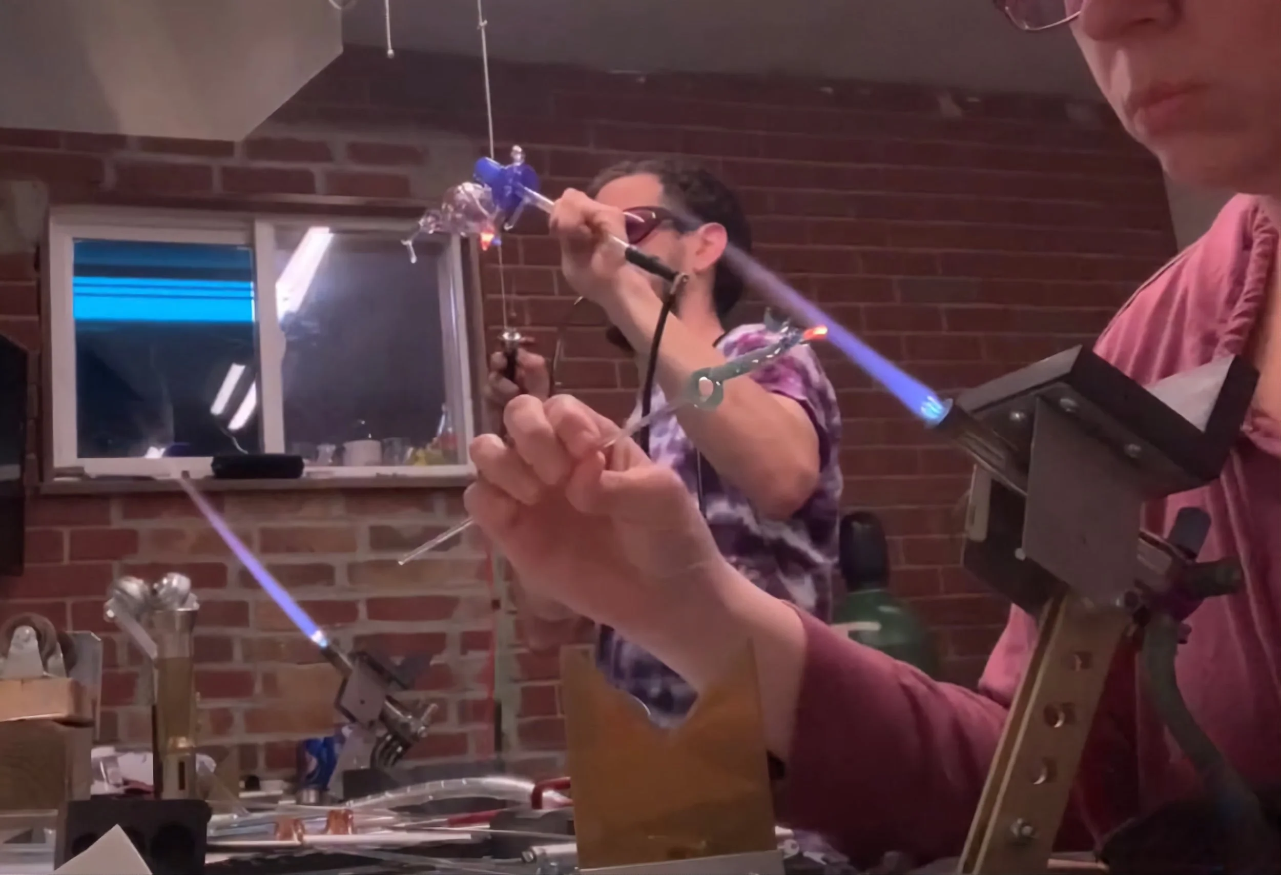 Two people working with glassblowing equipment; both of them is using a torch to shape glass while inside a room with brick walls.