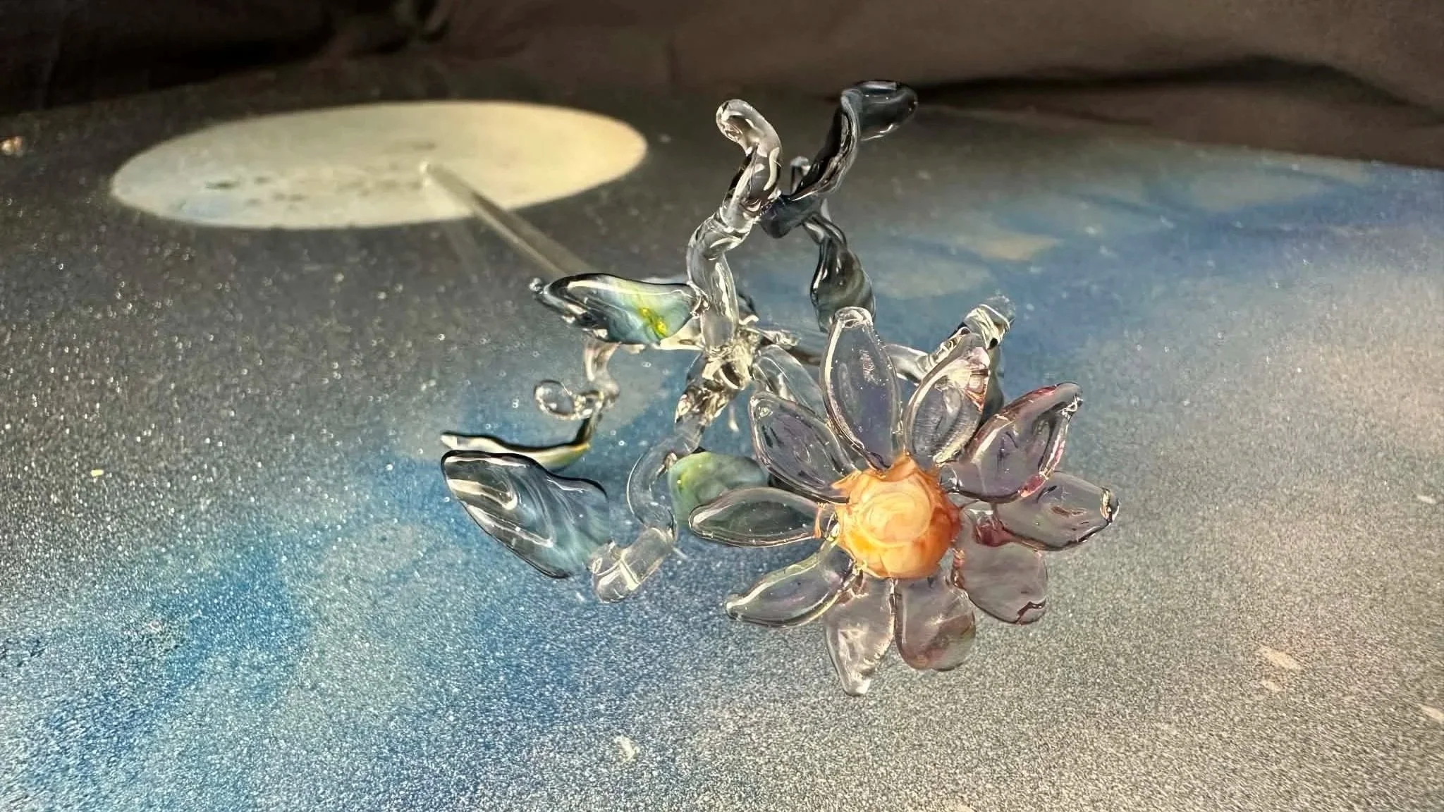 Glass flower sculpture with a glowing center, sitting on a textured dark surface with spotlights reflecting off it.