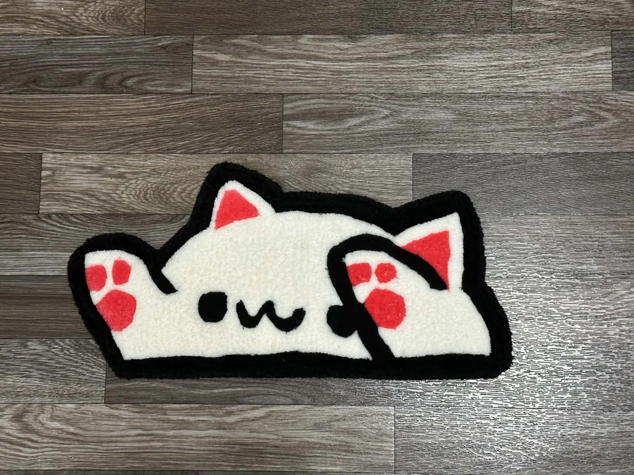 A fluffy rug designed to look like a cartoon cat's face with white, black, and red colors, placed on a dark wood floor.