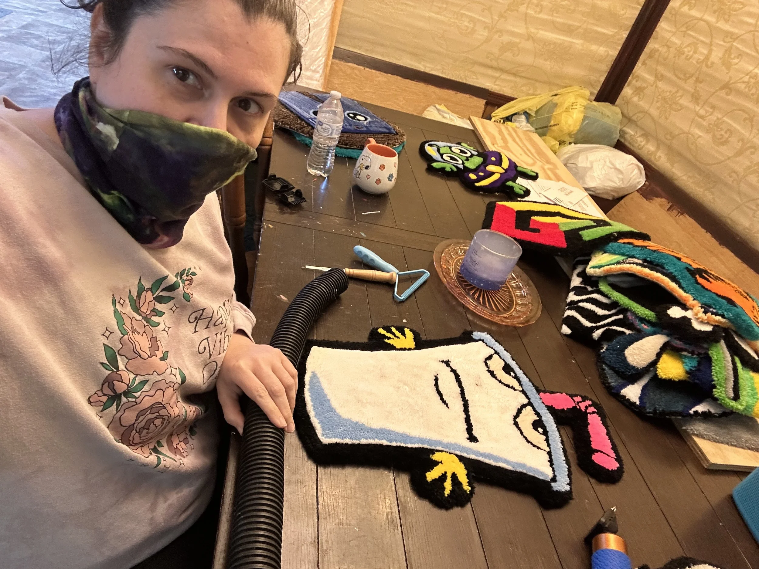 A woman with dark hair tied back, wearing a pink hoodie with floral design, sitting at a table covered with cats-themed and colorful craft items, including fuzzy cat-shaped rugs, a water bottle, a mug, and craft tools in a room with yellowish patterned walls.