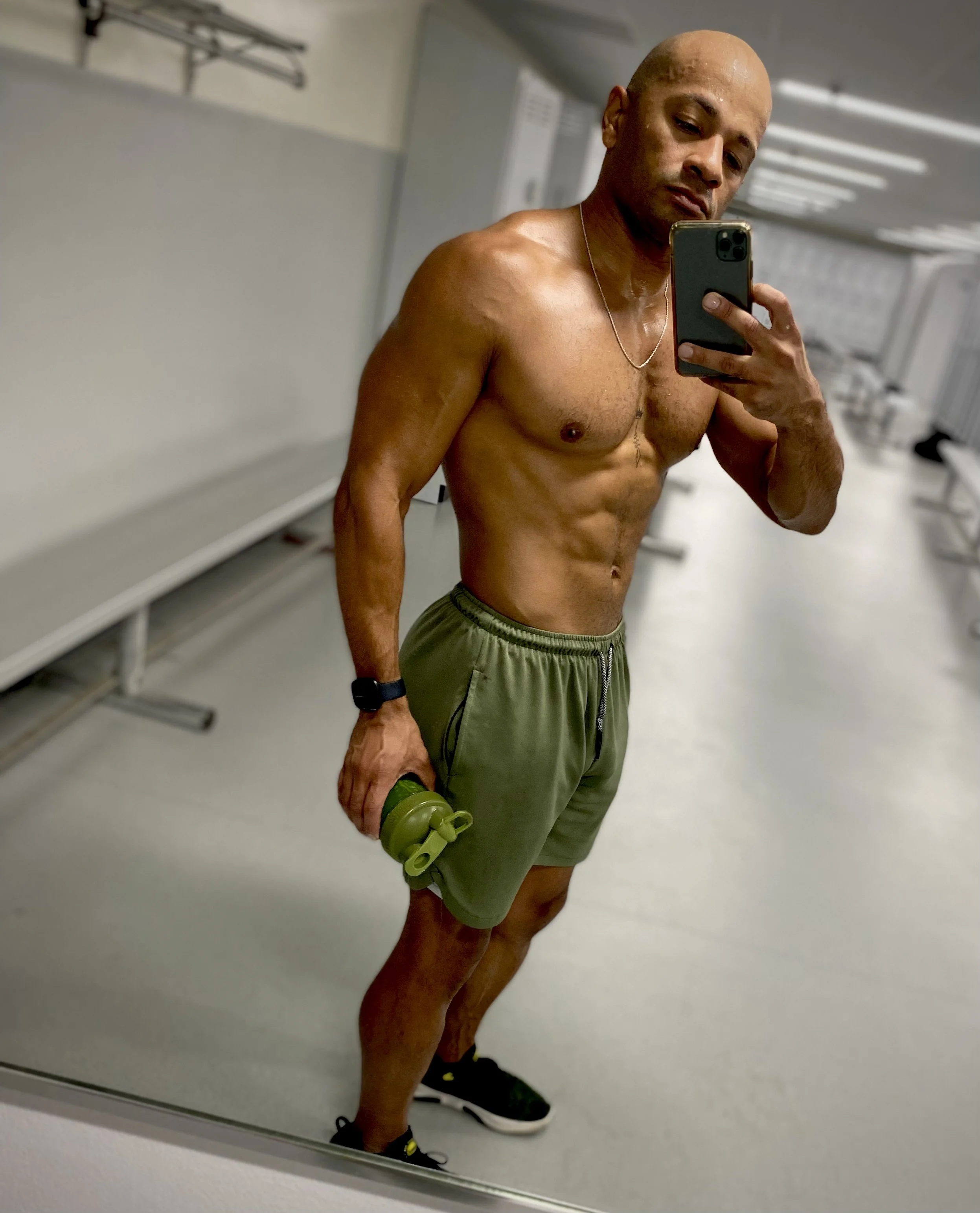A shirtless man wearing green shorts taking a selfie in a gym locker room mirror, holding a green water bottle, with a fitness tracker on his wrist. UnalomeFitness. Coach Bobby.