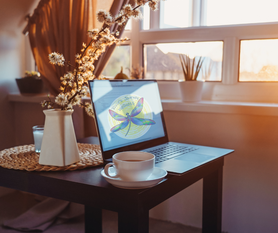 A cozy home workspace with a laptop, a white cup on a saucer, a glass of water, and a vase with dried flowers on a wooden table near a window during sunset.