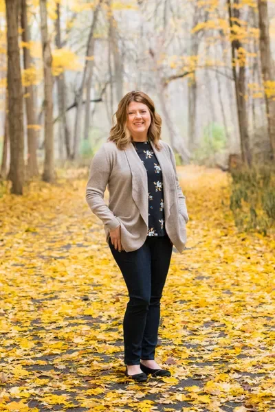 A woman standing on a leaf-covered trail in a forest during fall, smiling, wearing a beige blazer, floral blouse, dark jeans, and black shoes.