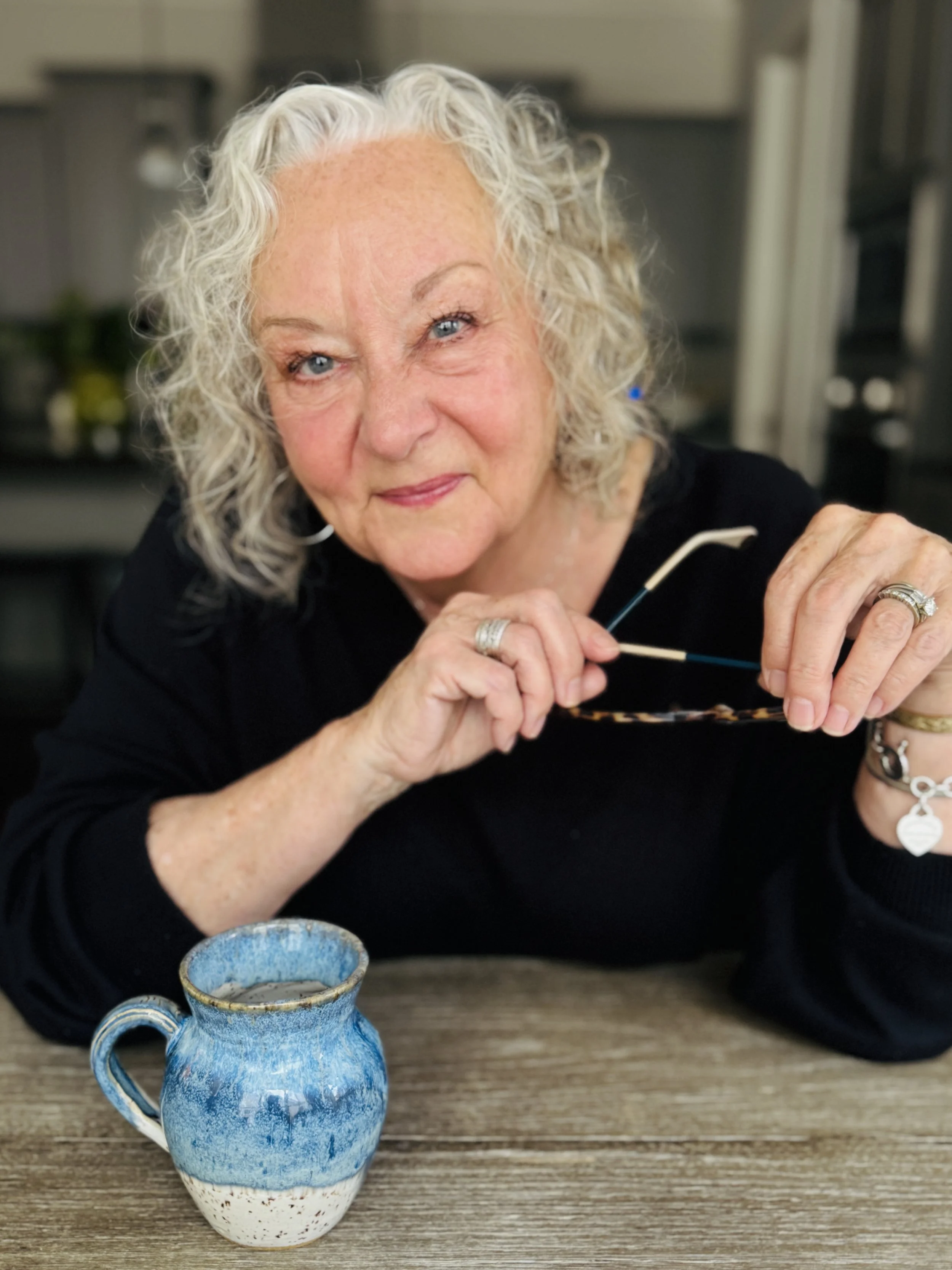An elderly woman with curly gray hair and blue eyes, wearing a black top, holding glasses, sitting at a wooden table with a blue ceramic mug in front of her.