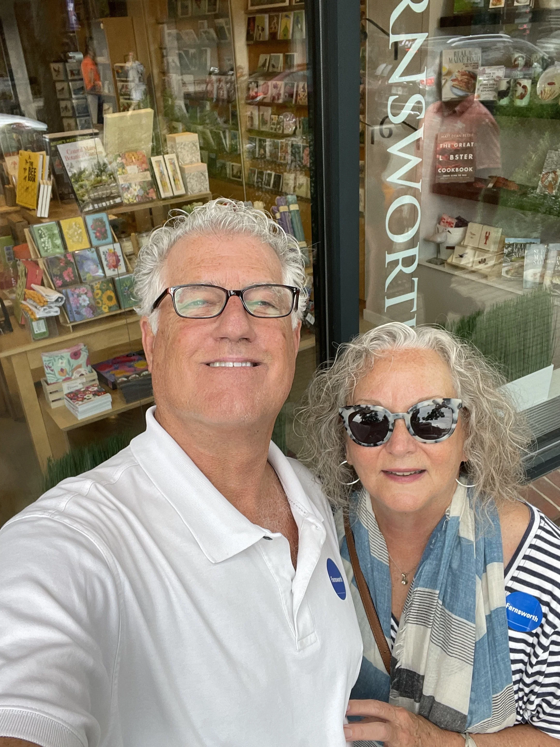 A photo of two older individuals taking a selfie outside a store with books and greeting cards visible through the window. The man is smiling and wearing glasses and a white polo shirt, while the woman is wearing sunglasses, a striped shirt, and a beige scarf.