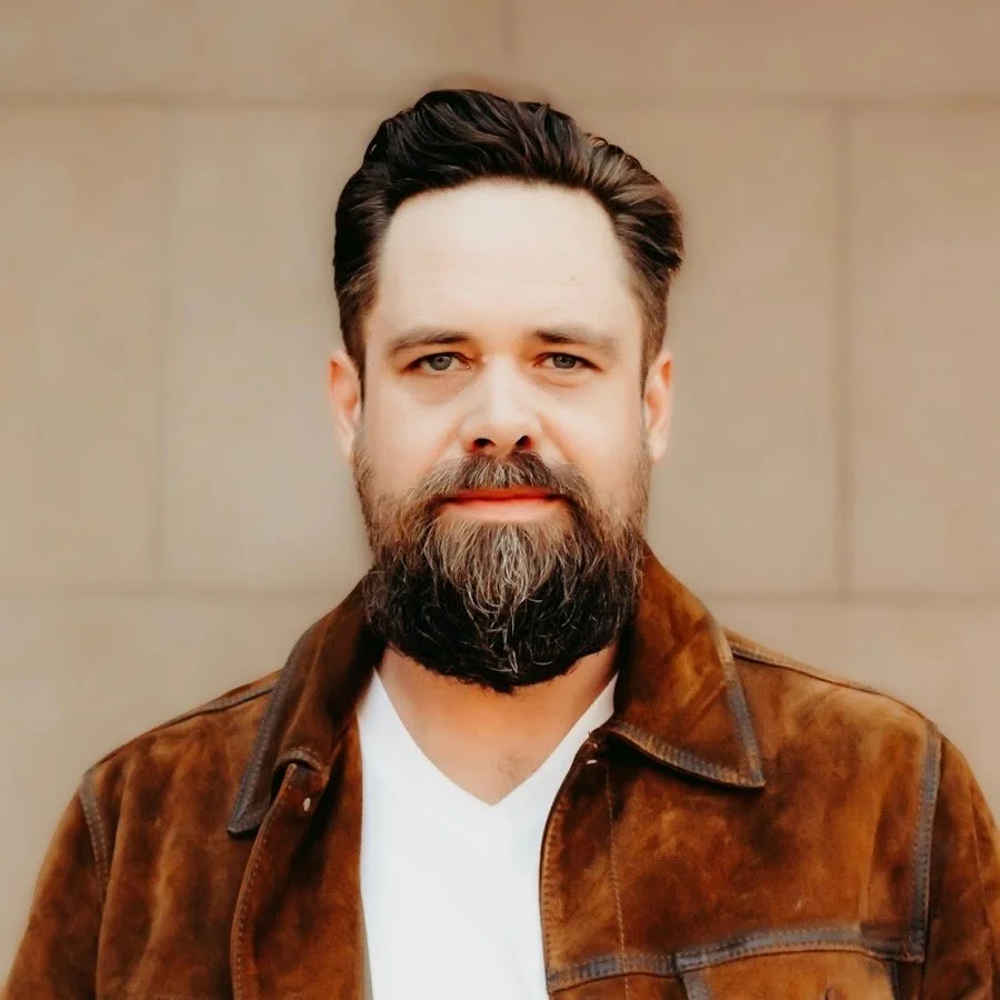A smiling man with a beard wearing a brown jacket against a concrete wall..