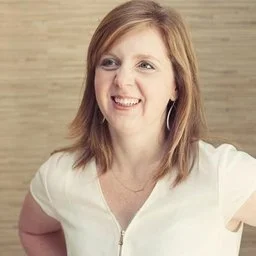 A woman with shoulder-length brown hair smiling, wearing a white top, standing against a beige wall.