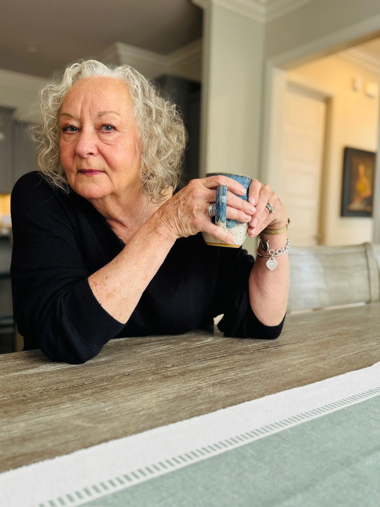 An elderly woman with curly gray hair sitting at a wooden table, holding a ceramic mug with a blue handle, wearing a black top and jewelry, in a well-lit room with framed artwork on the wall.