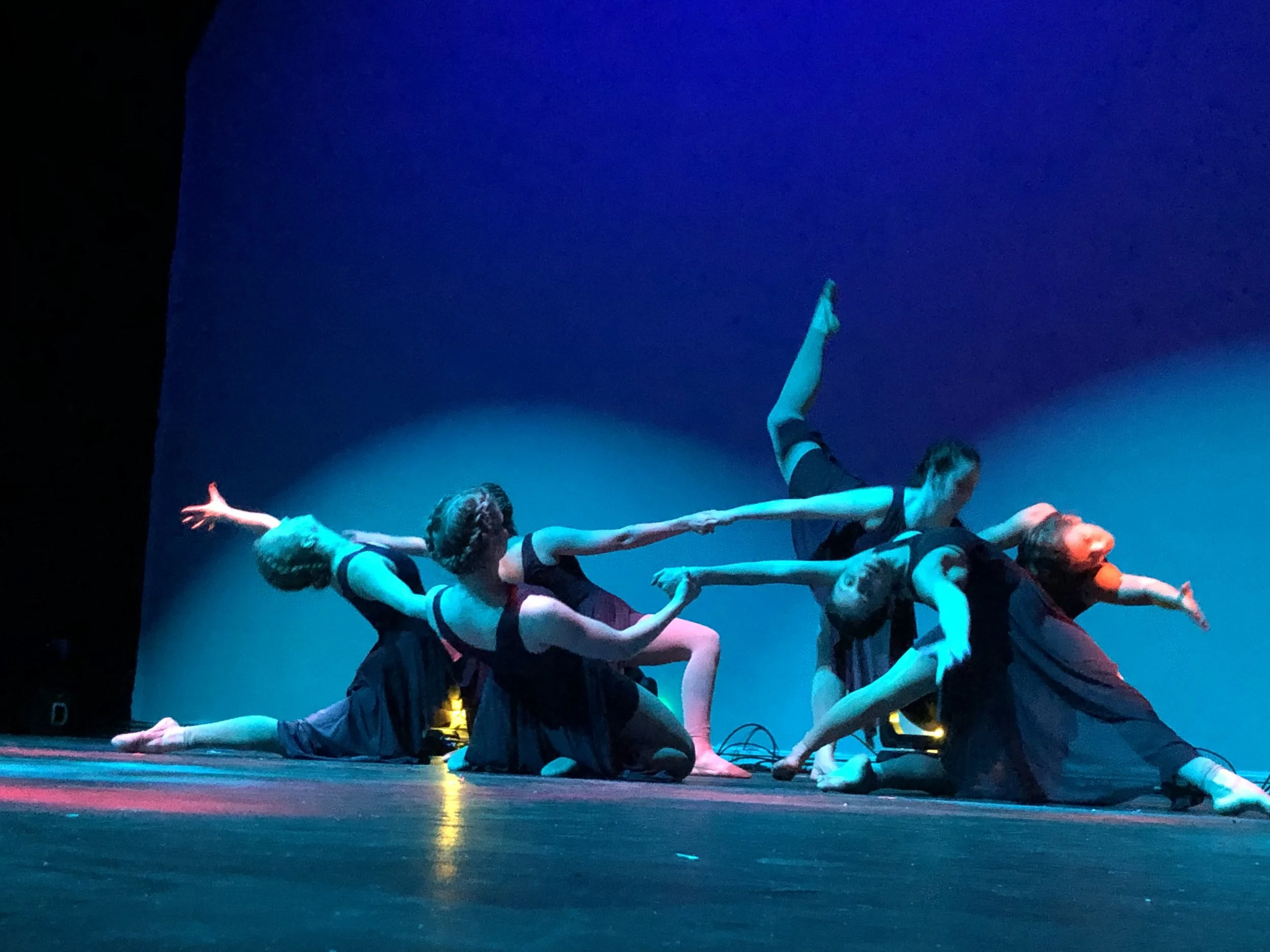 Group of dancers performing on stage, some lying on the floor while others are supported by their hands and feet, with a dark background and colorful stage lighting.