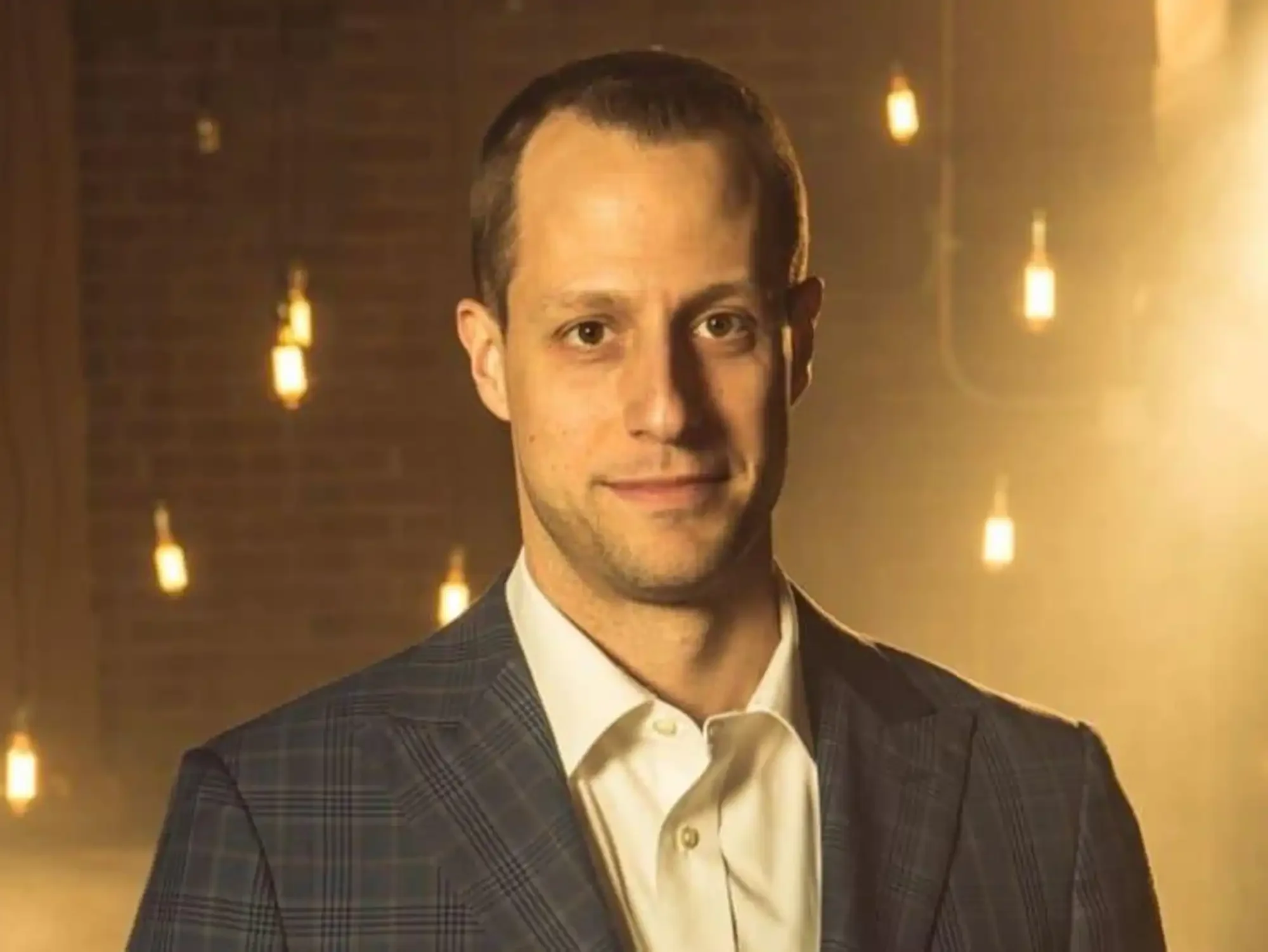 A man with short dark hair wearing a checked suit, white shirt, and tie, smiling slightly, standing in front of a warm, glowing background with hanging lights.