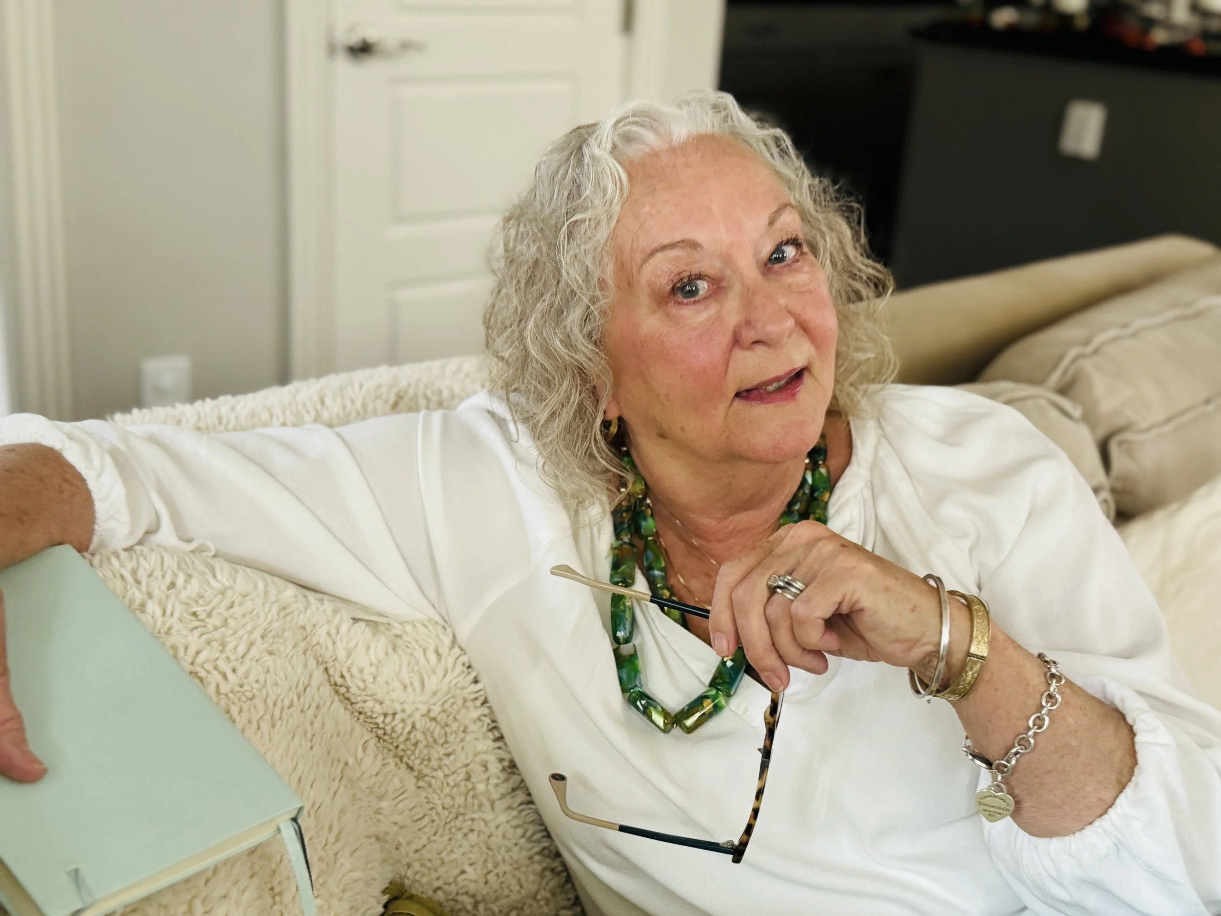 An elderly woman with curly gray hair, blue eyes, and wearing jewelry, sitting on a cream-colored couch in a room with beige walls, smiling and holding another person's hand.
