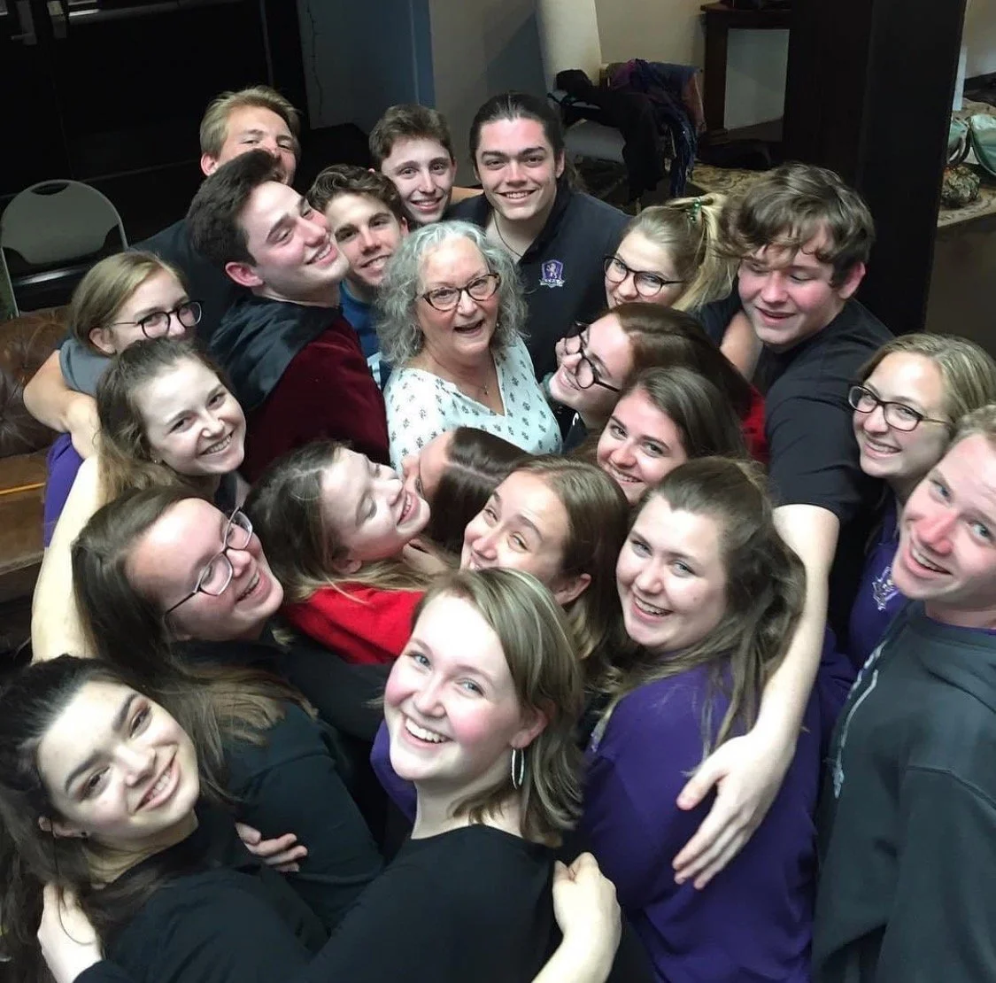 A group of teenagers and an older woman are hugging and smiling for a group photo in an indoor setting.