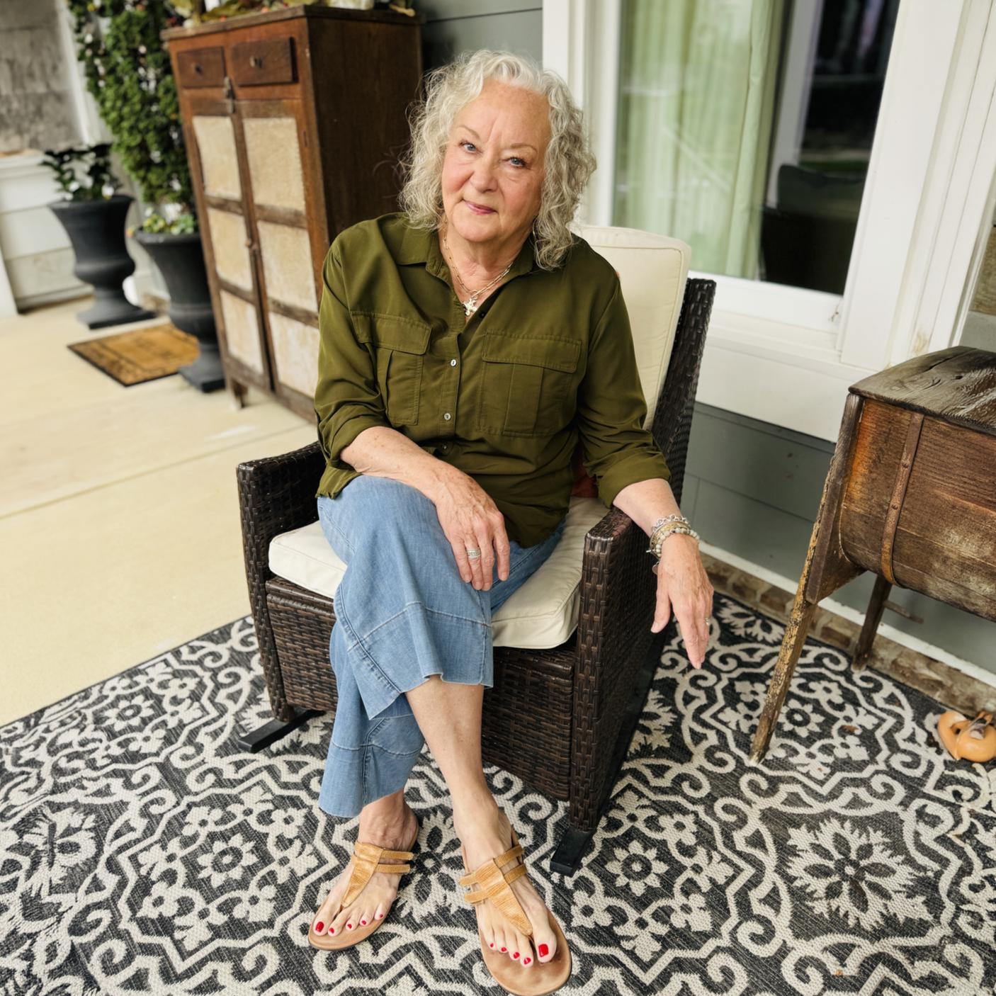 An elderly woman with curly gray hair sitting on a wicker chair on a porch or patio, wearing a green shirt, blue jeans, and tan sandals with red toenail polish, smiling slightly.