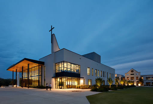 Modern church building with a cross on top, large glass windows, and a lit entrance at dusk.