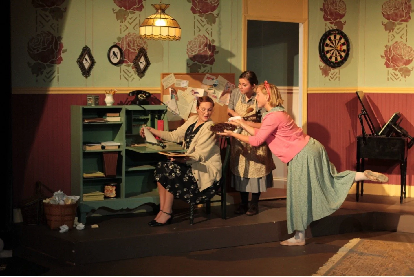 Scene from a play showing three women in a 1950s-style kitchen with floral wallpaper. One woman is sitting at a desk with a typewriter, another is standing nearby with a pie, and the third is balancing on one leg while offering the pie, all engaged in a humorous moment.
