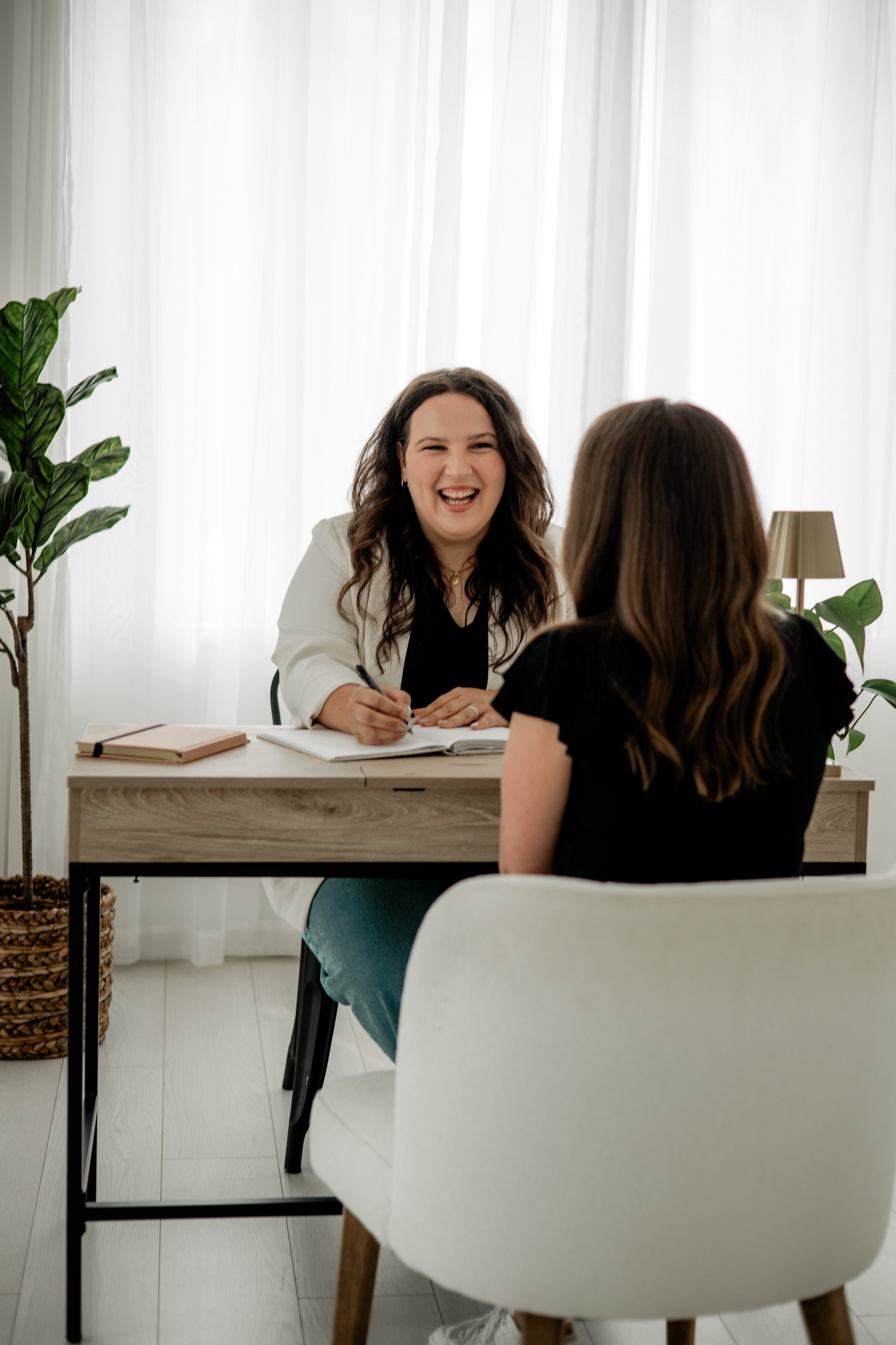 A woman with long dark hair and a white blazer smiling and writing notes during an interview or consultation with another woman sitting across from her, in a bright room with sheer curtains and plants.