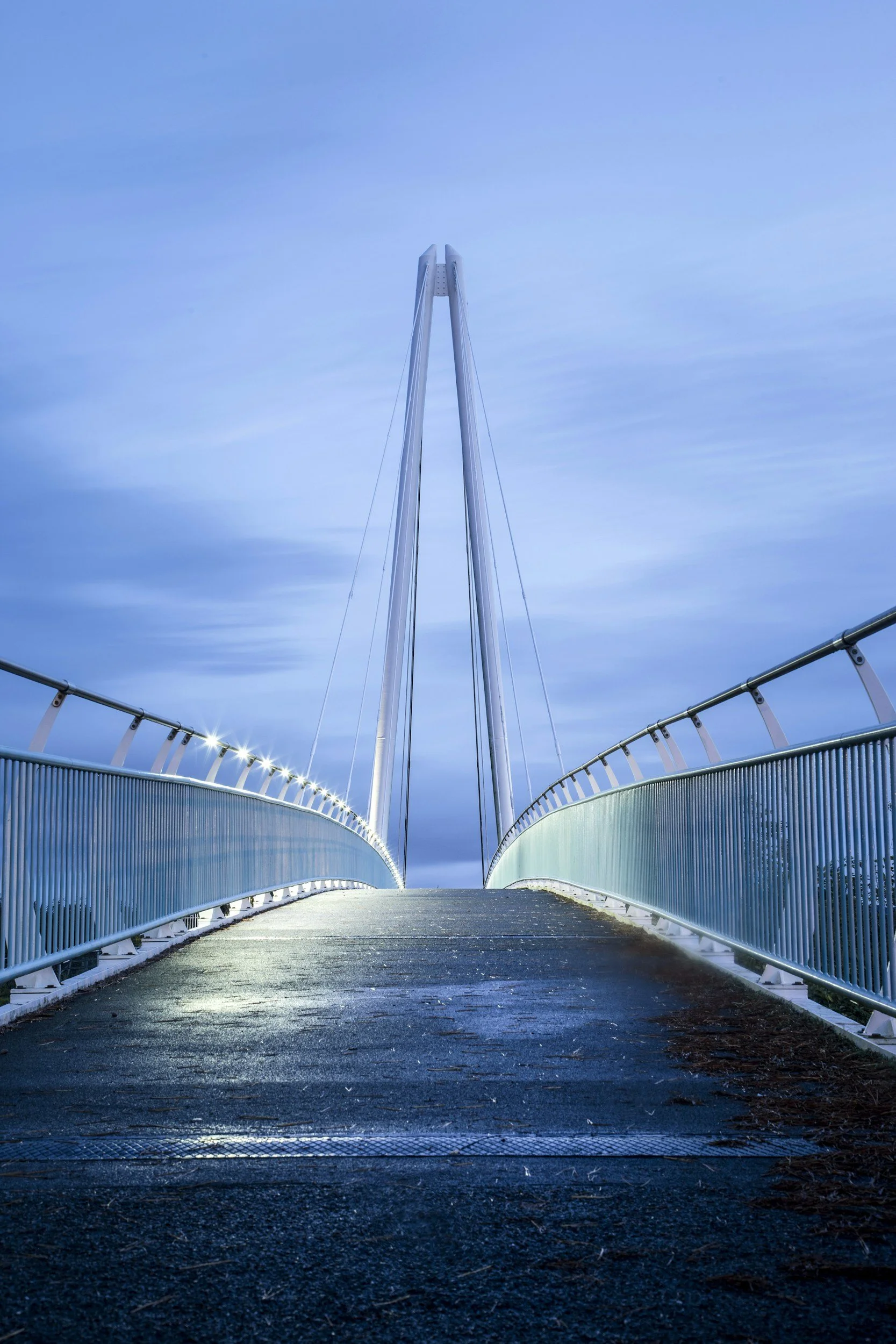 A modern cable-stayed pedestrian bridge with a sleek, white, high-arch design against a cloudy blue sky, illuminated by lights along the sides.