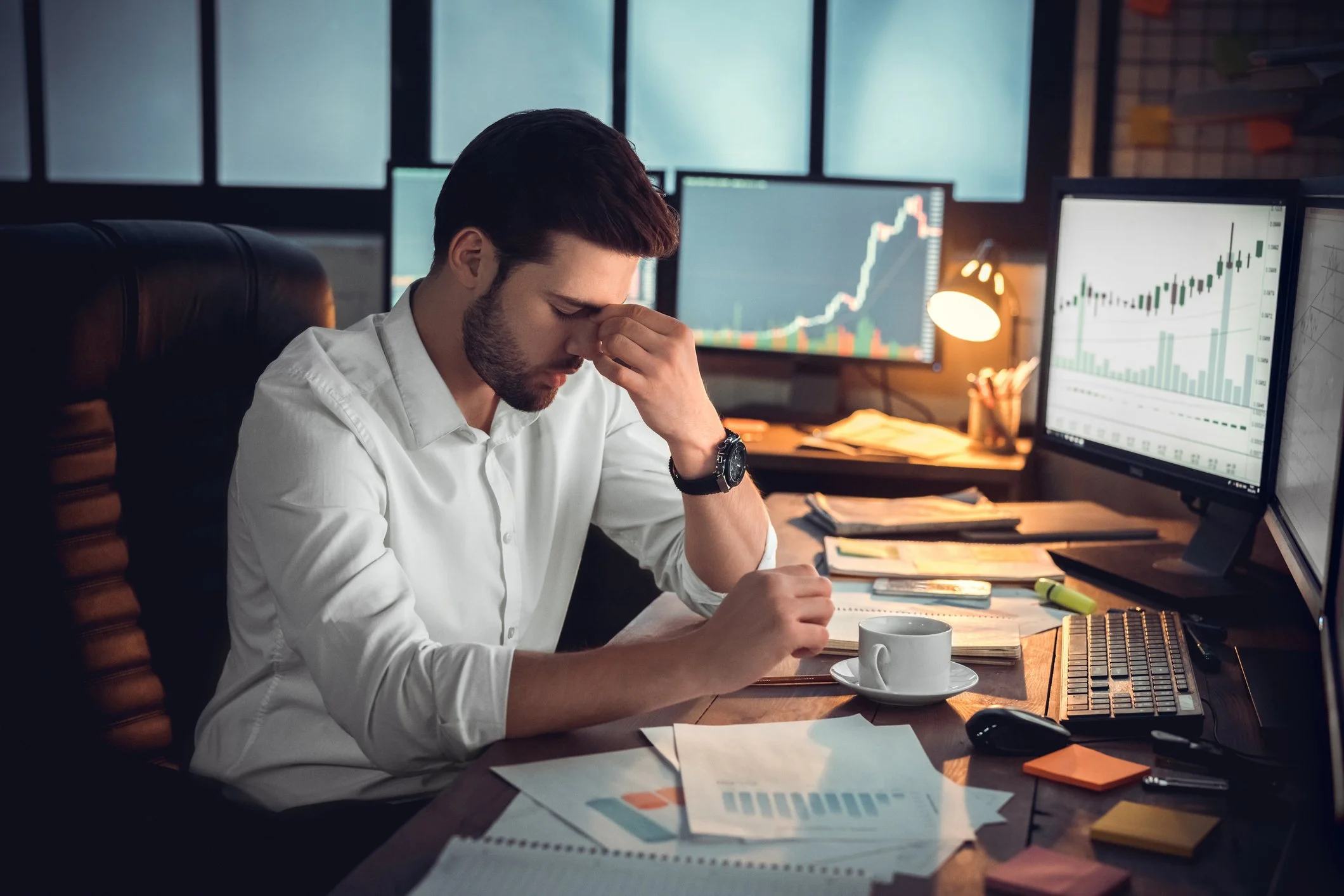 An upset man sitting at a cluttered office desk with financial documents, a coffee cup, and multiple computer monitors displaying stock charts, holding his head with one hand.
