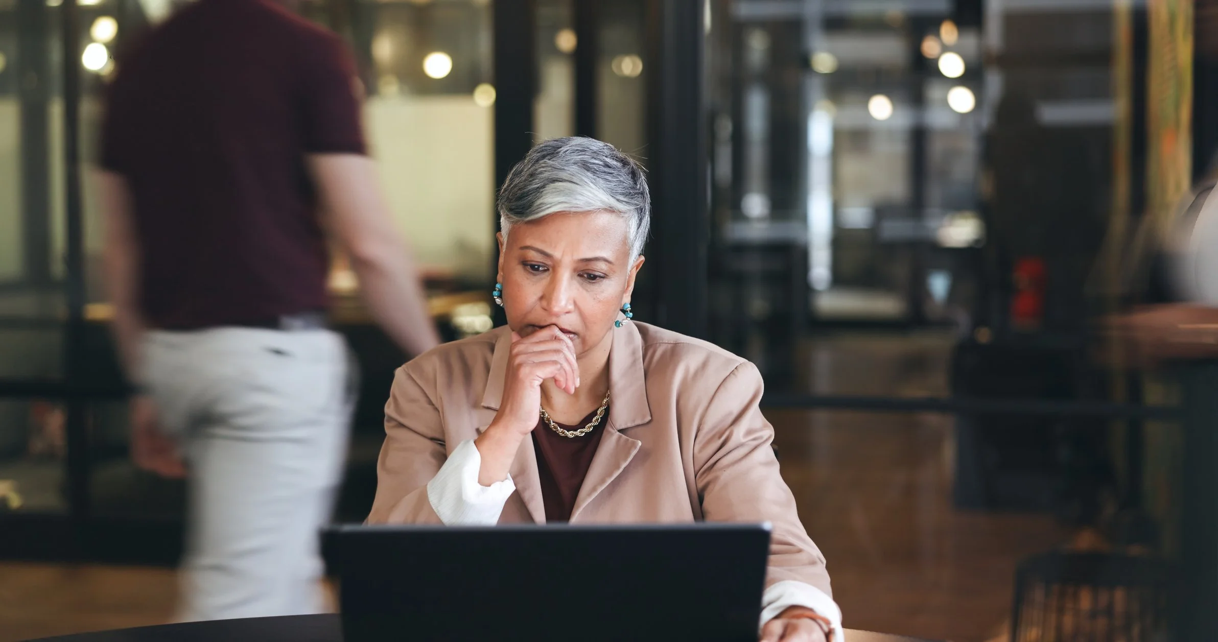 An older woman with short gray hair and earrings is sitting at a table, looking at a laptop with a concerned or thoughtful expression.