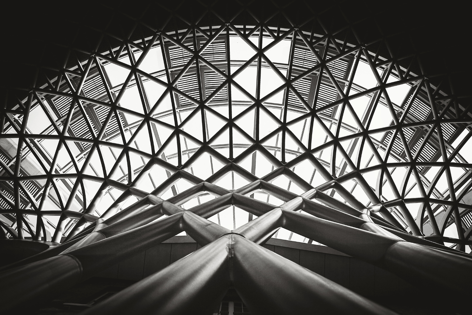 Black and white view looking up at the intricate lattice structure of a modern architectural ceiling with intersecting beams and exposed steel supports.