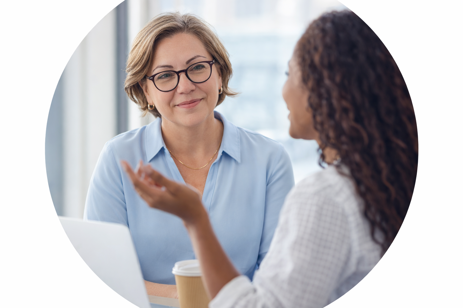 Two women engaged in a conversation indoors, one with short blond hair and glasses, and the other with long curly hair, with a coffee cup and laptop on the table.