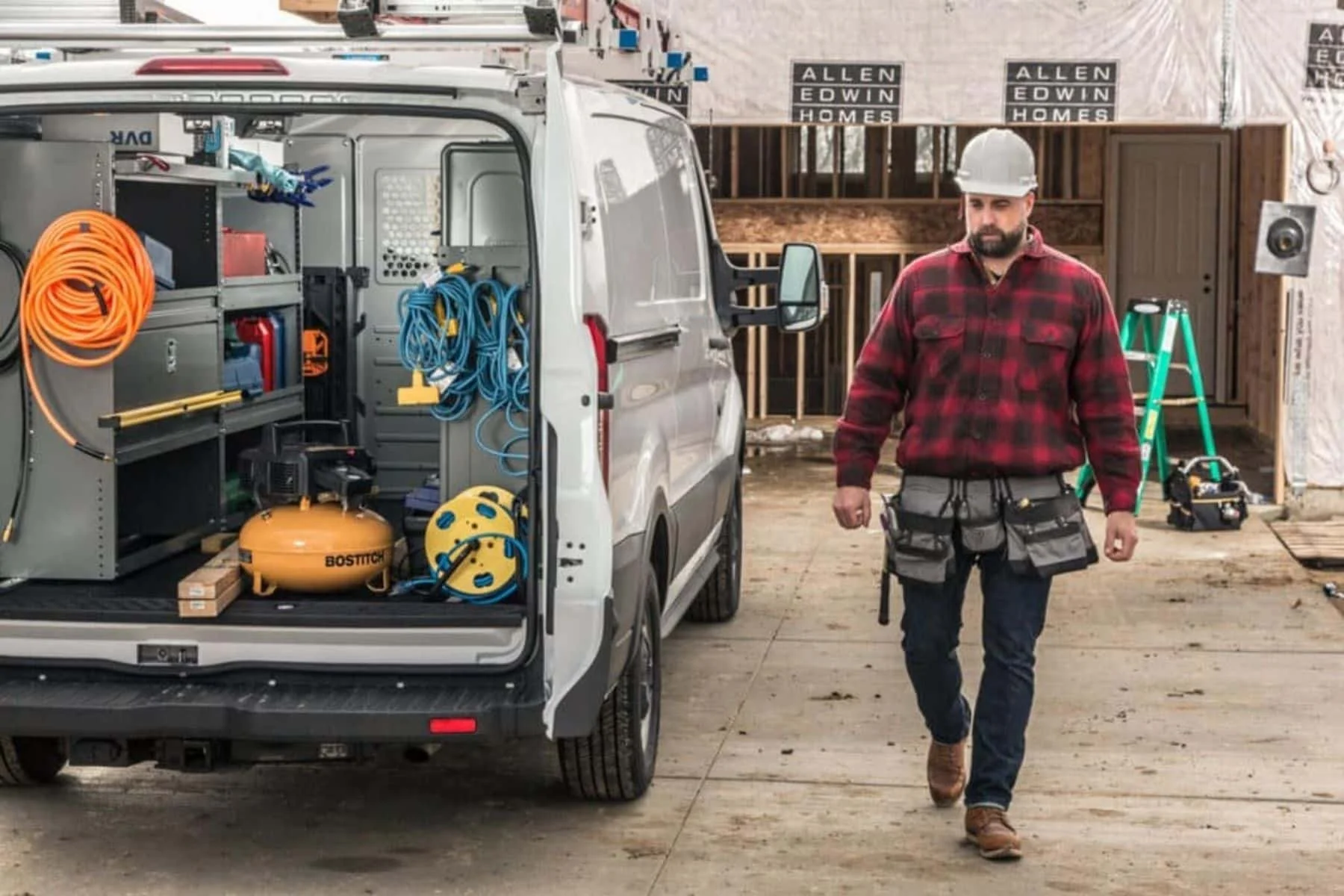 A construction worker wearing a gray hard hat and red plaid shirt walking near an open work van with equipment and tools inside, at a building site under construction.