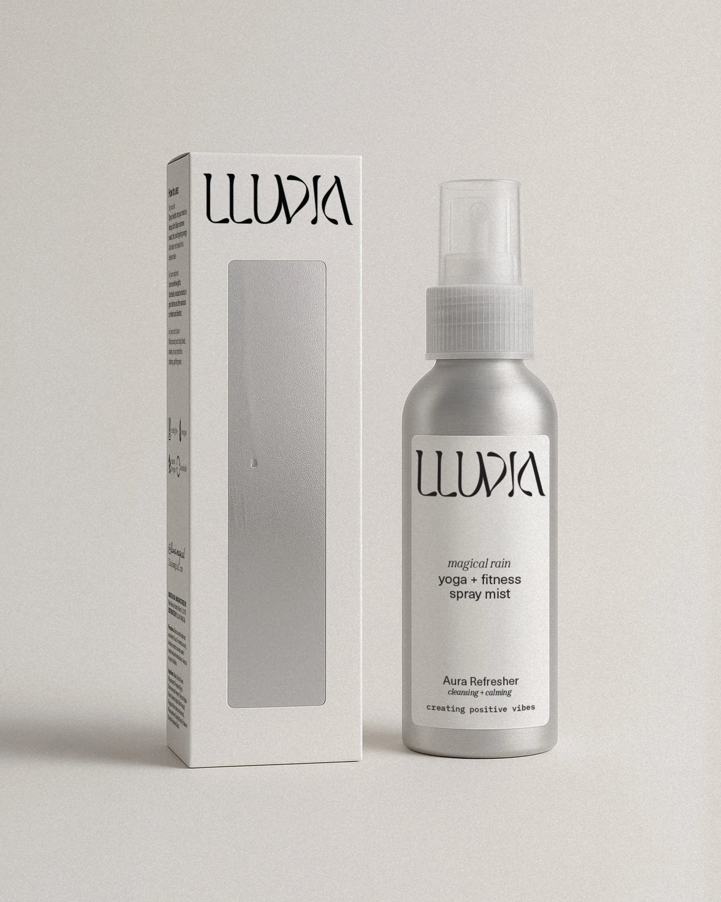 A white spray mist bottle labeled 'Lluvia' with 'magical rain yoga + fitness spray mist' and 'Aura Refresher cleansing + calming' next to a matching boxed product against a light gray background.