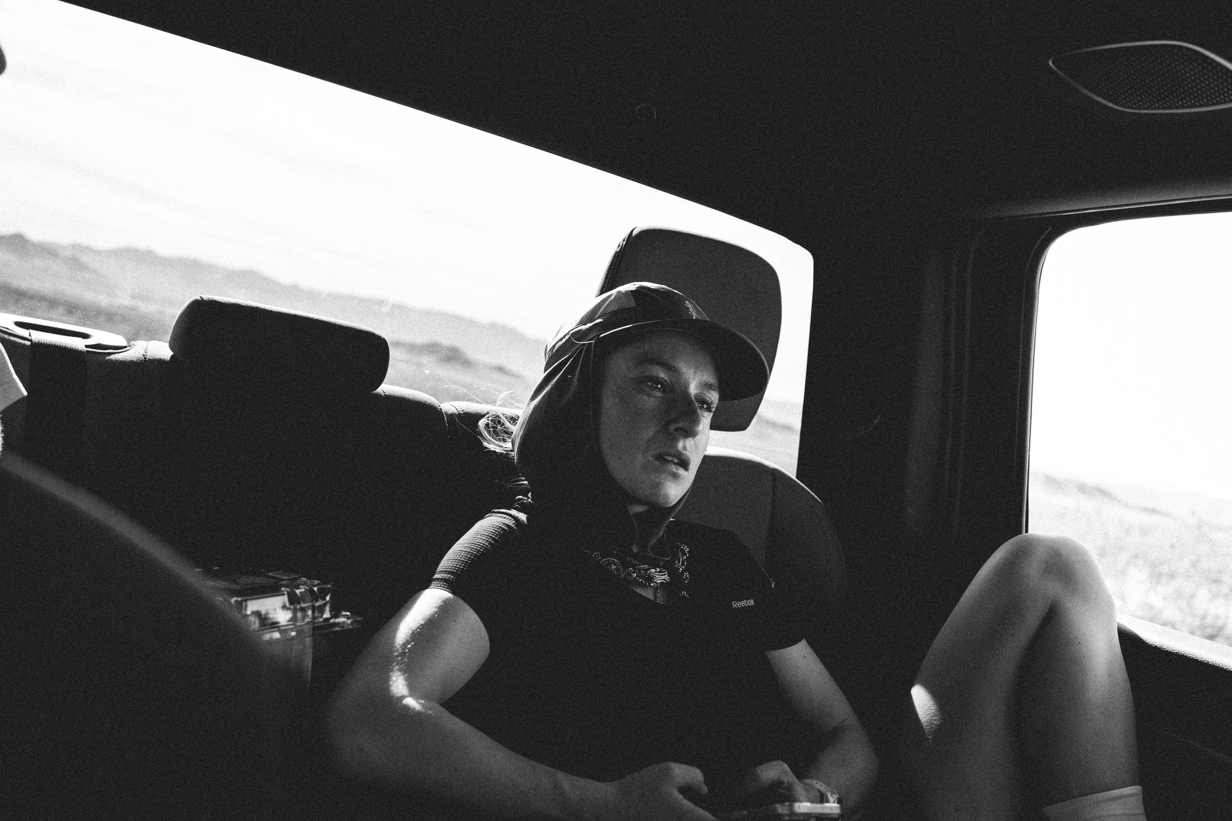 A black-and-white photo of a person sitting in the backseat of a vehicle, wearing a cap and casual clothes, looking outside through the window at a landscape of open land and mountains in the distance.