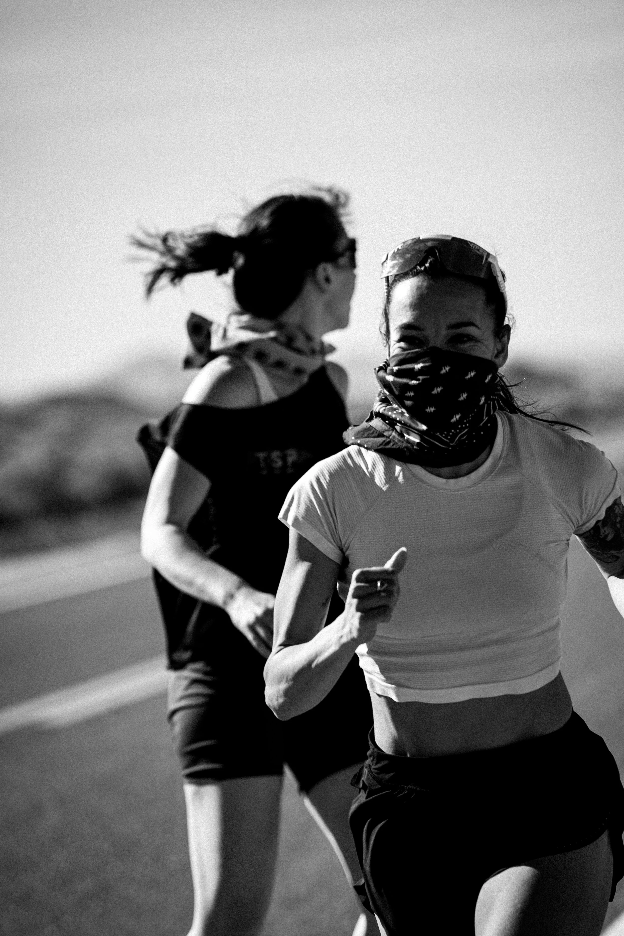 Black and white photo of two women running outdoors, with the woman in the foreground smiling and wearing a face covering, sportswear, and sunglasses on her head.
