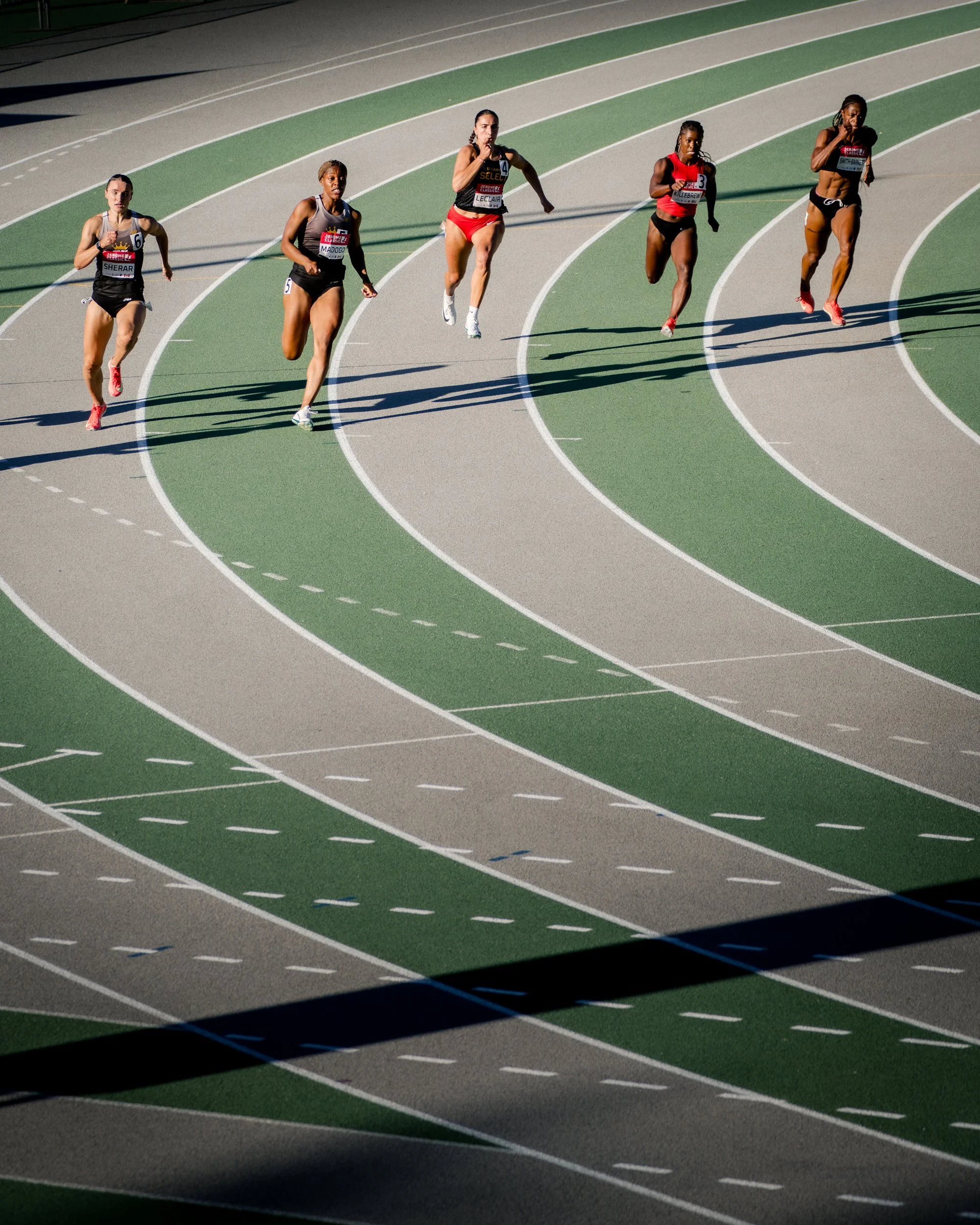 Five female athletes running on a curved outdoor track in a race, wearing athletic gear and numbered bibs.
