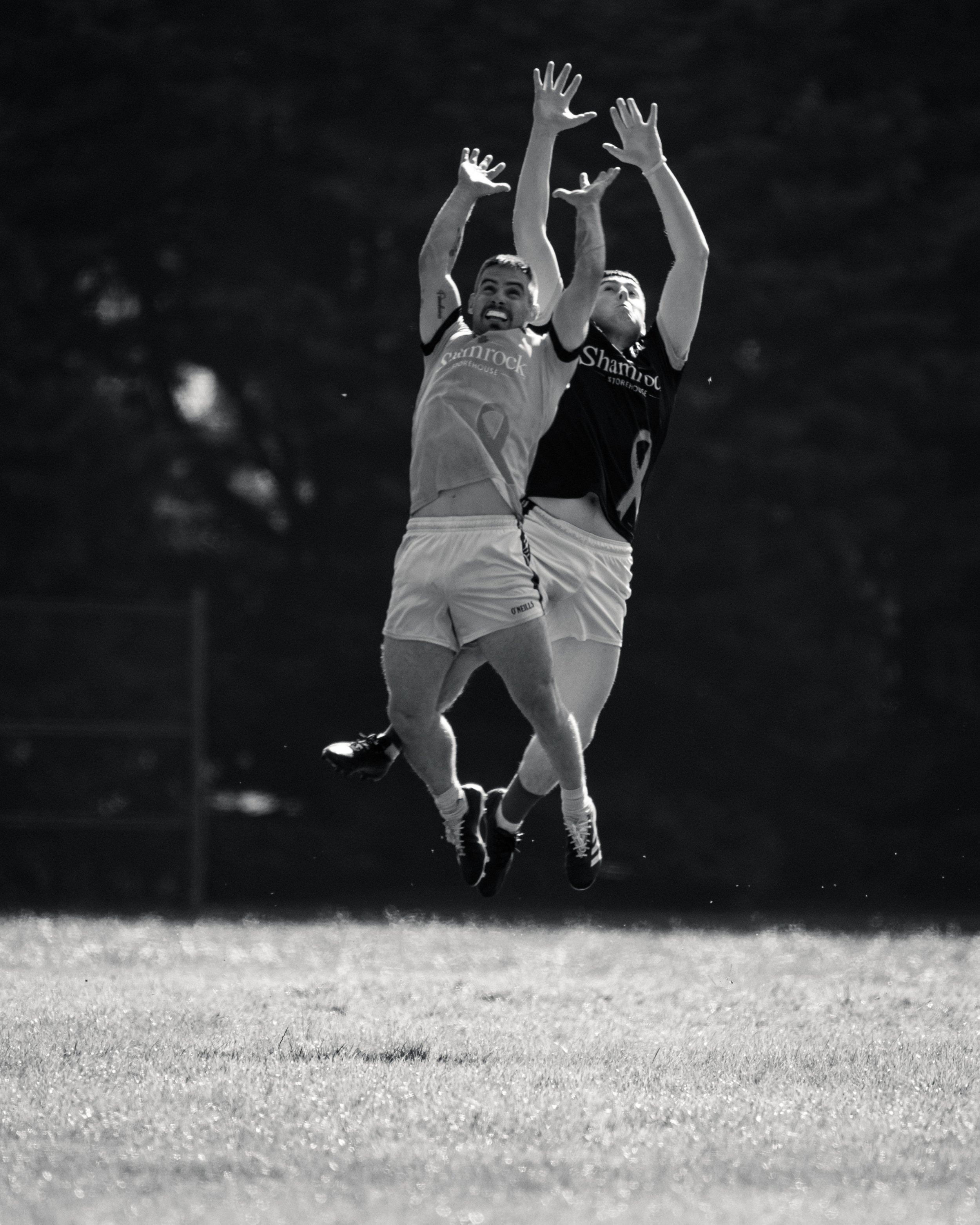 Two soccer players jumping for a ball on a grassy field, one in a dark jersey and the other in a light jersey, during a game.