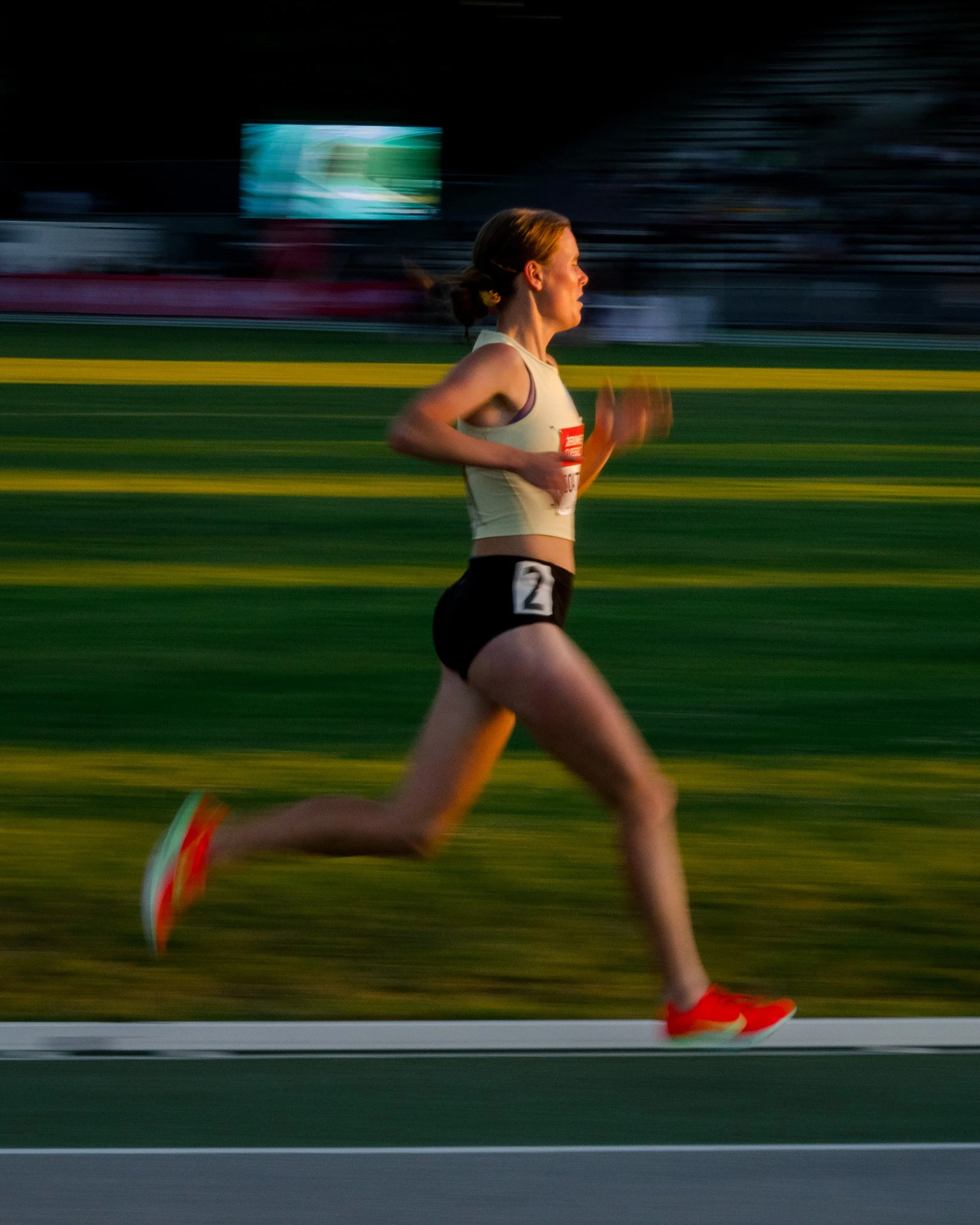 A female runner in a race, wearing a light-colored tank top, black shorts with a number 2, and red shoes, running on a track at dusk.