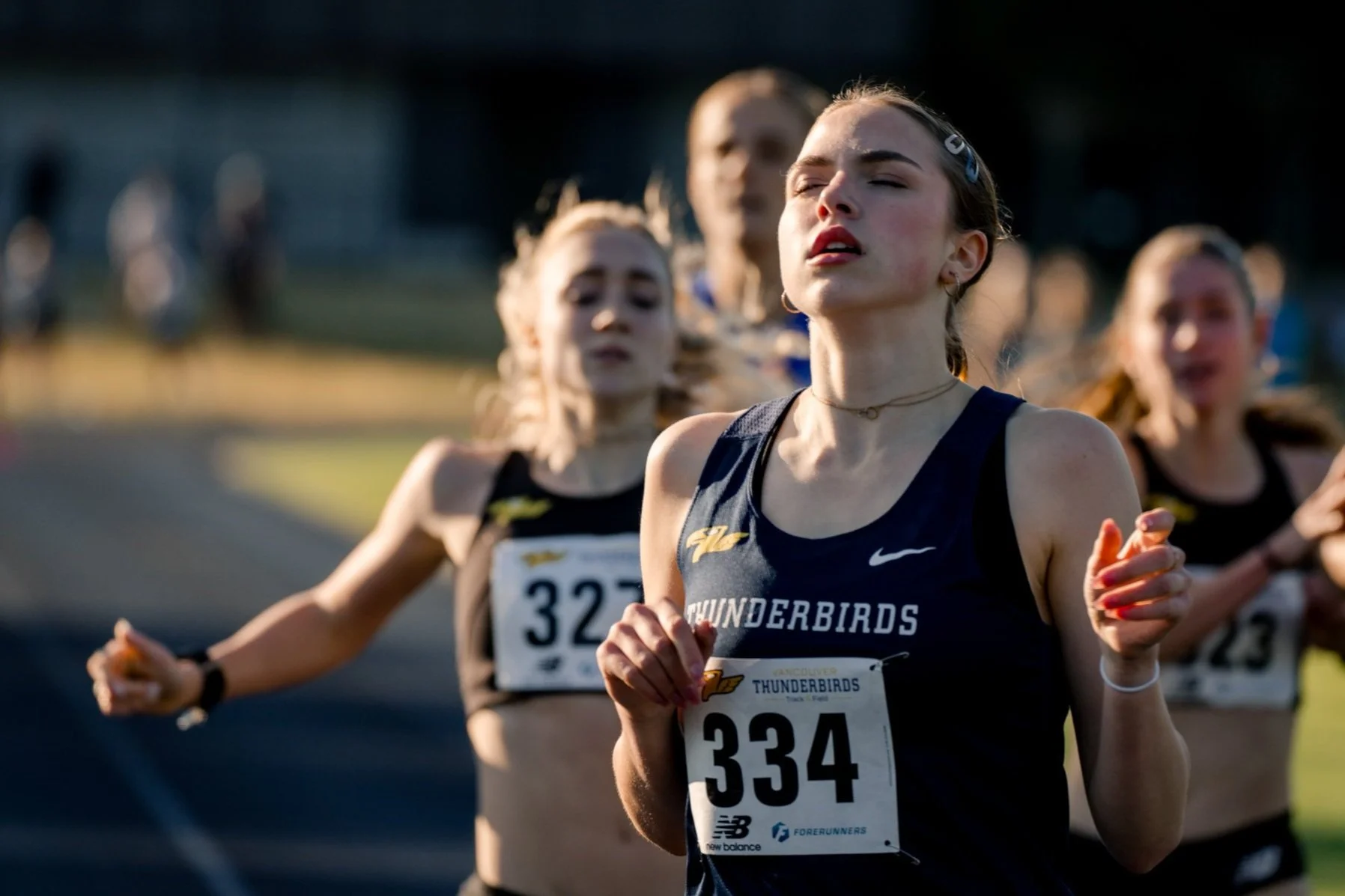 Women running in a race, wearing black athletic uniforms with 'Thunderbirds' printed on the front, race bibs with numbers 334, 327, and 329, on a track field during sunlight.
