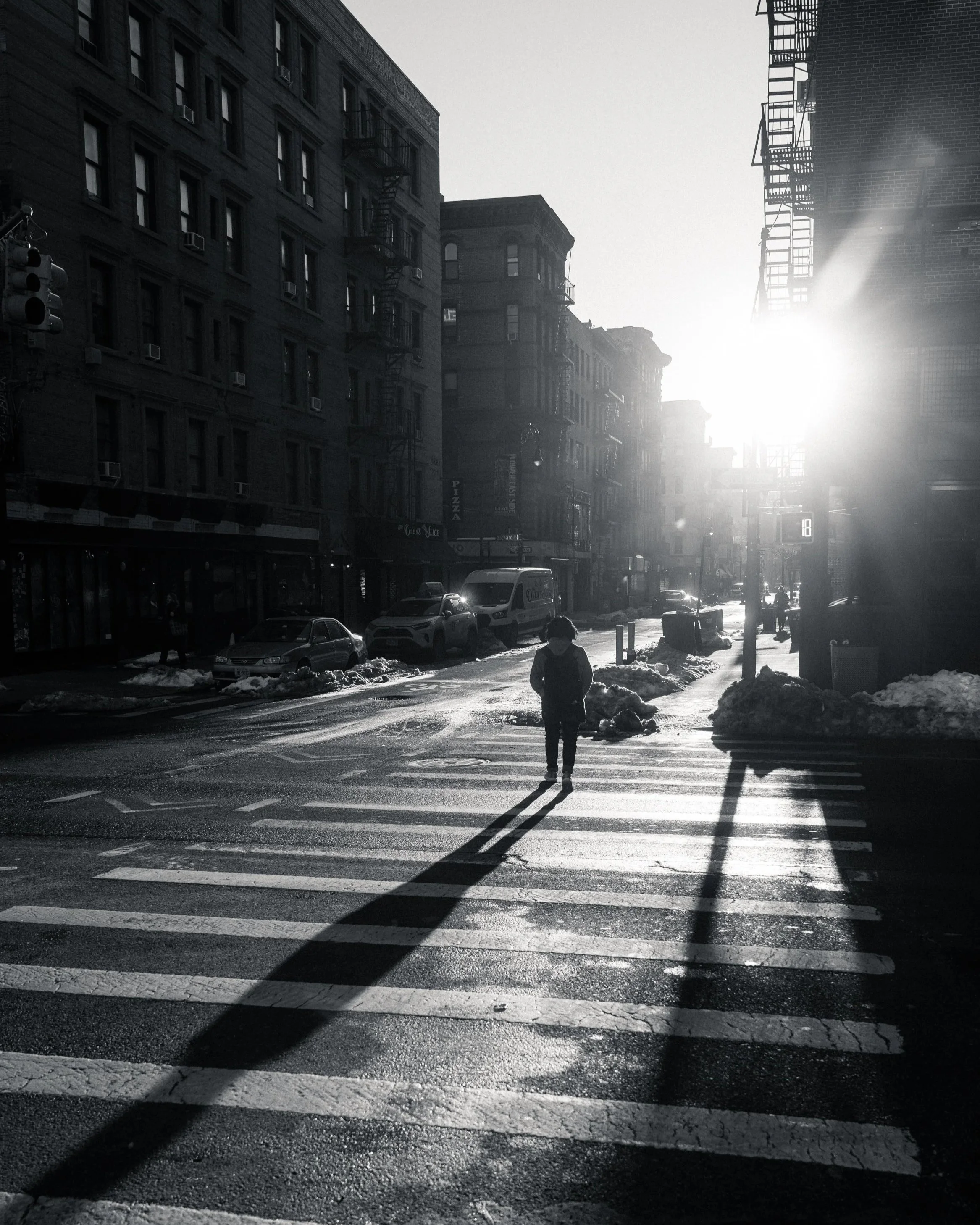 City street scene during sunset with buildings, parked cars, snow on the sidewalk, and a person walking across a crosswalk, casting a long shadow.