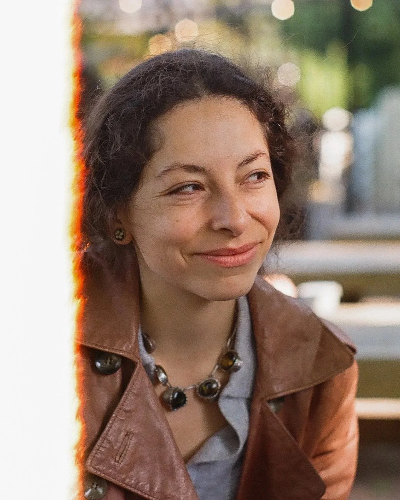 A woman with short, curly dark hair wearing a brown leather jacket and a black beaded necklace smiling outdoors with blurred lights and greenery in the background.