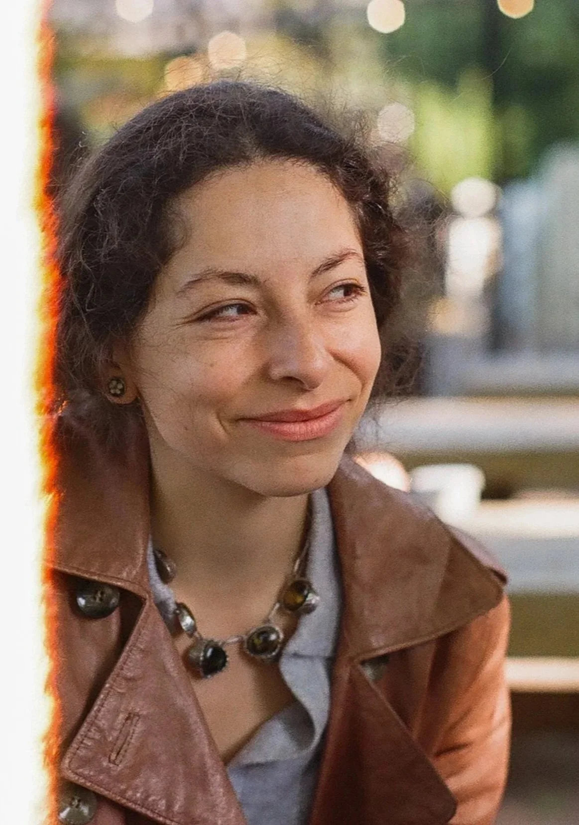 A woman with short, curly dark hair wearing a brown leather jacket and a black beaded necklace smiling outdoors with blurred lights and greenery in the background.