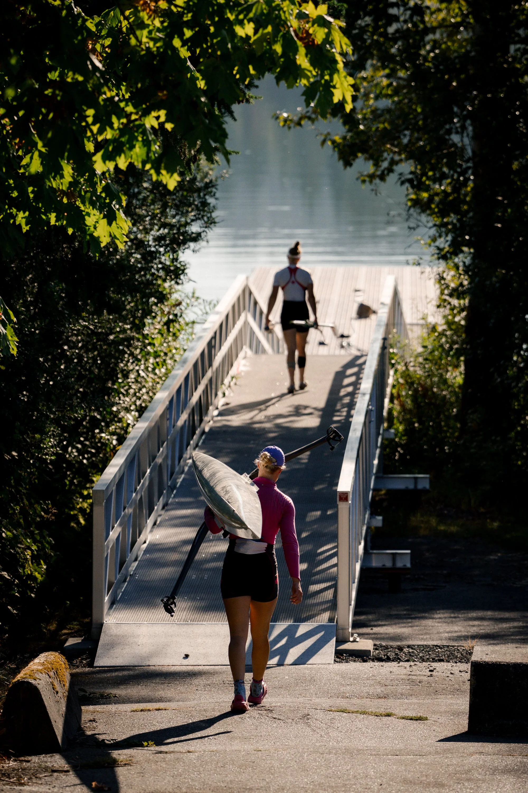 Two women walking down a ramp towards a body of water, one carrying a kayak on her shoulder and the other holding a paddle, surrounded by green trees.