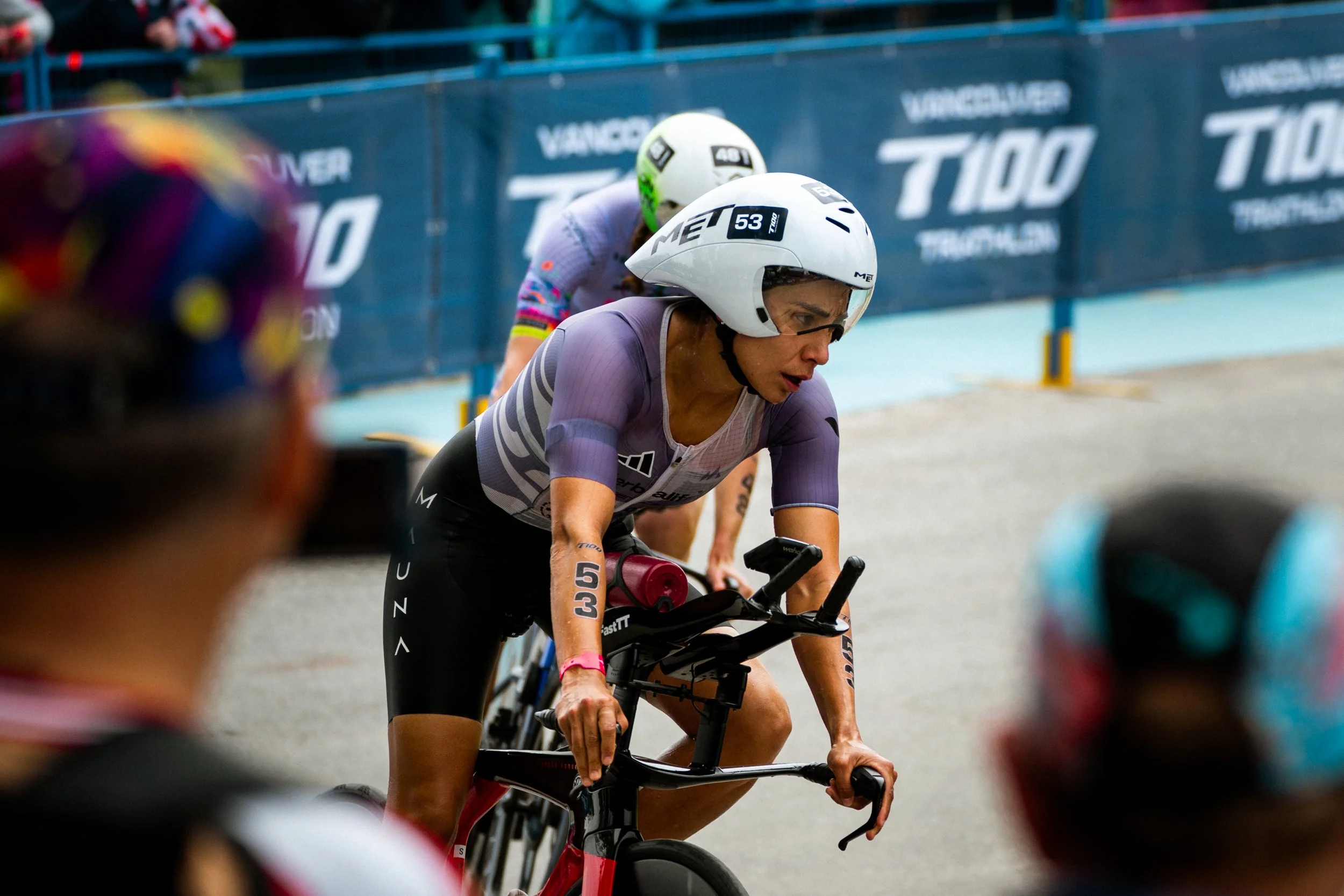 Athletes competing in a bike race, wearing helmets and racing gear, with a guardrail and advertising banner in the background.