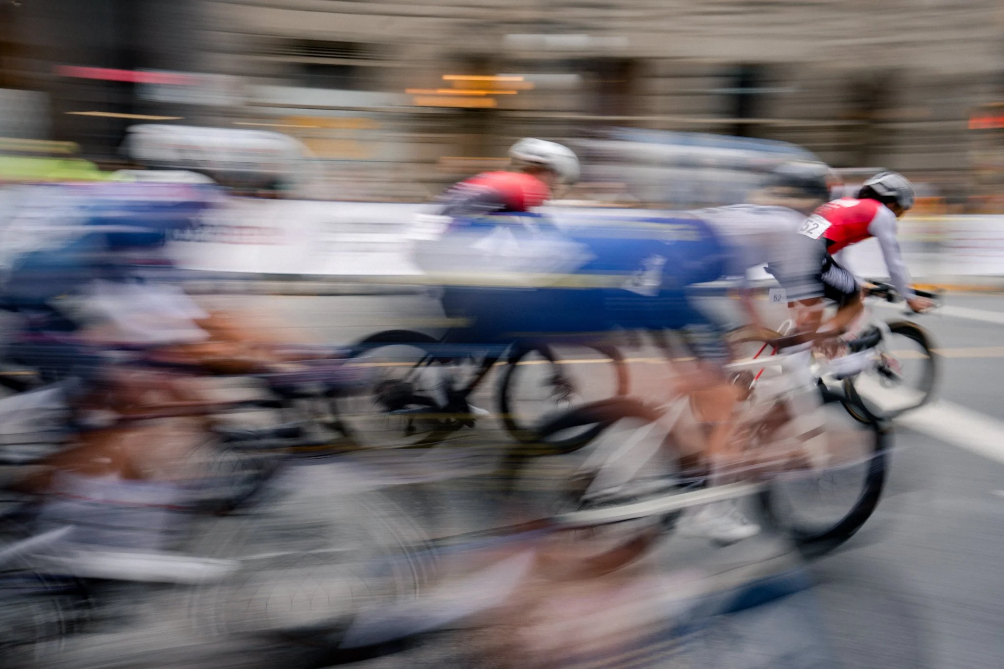 Blurred motion image of cyclists racing on a city street, with buildings and barriers in the background.