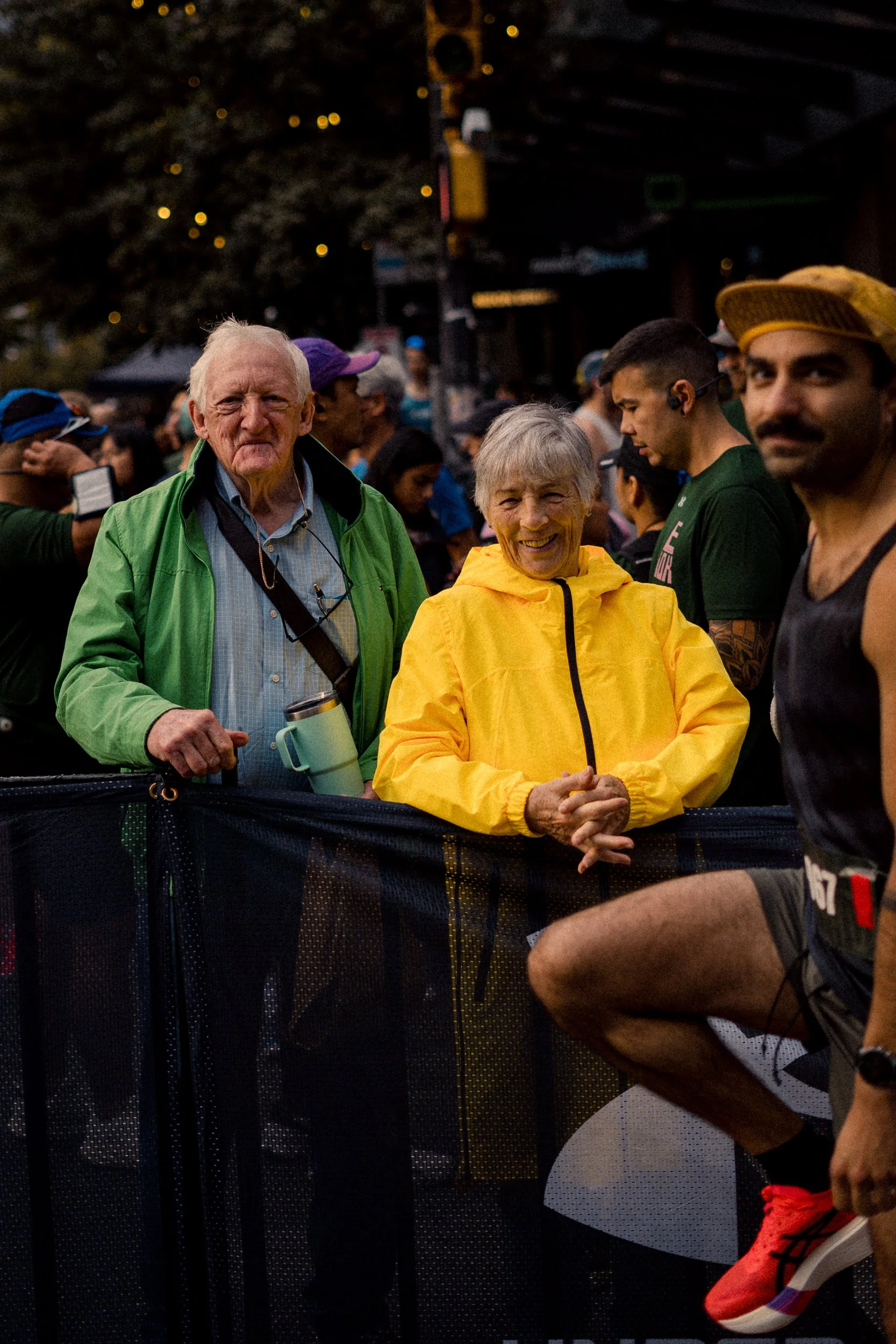 Crowd of people at an outdoor event, with two elderly individuals in the center smiling and wearing colorful jackets, surrounded by other attendees.
