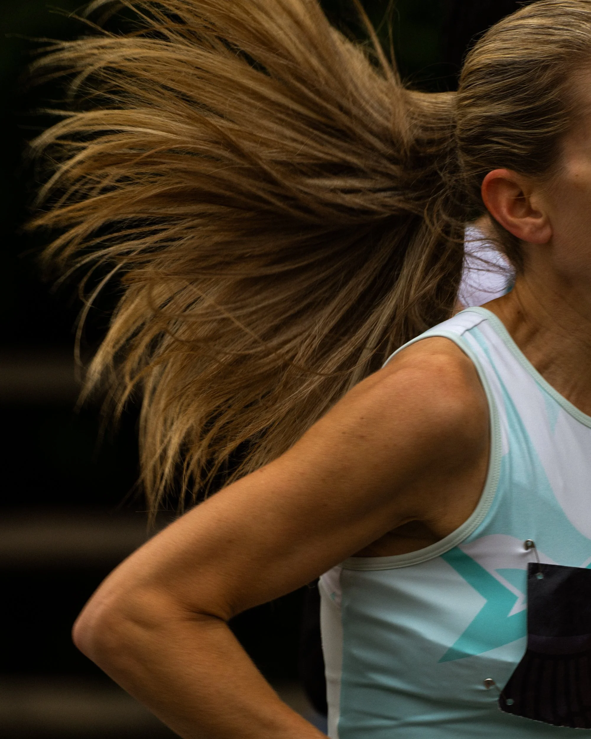 Close-up of a woman with long, blonde hair tied in a ponytail, wearing a light blue and white athletic tank top, with part of her face and shoulder visible.