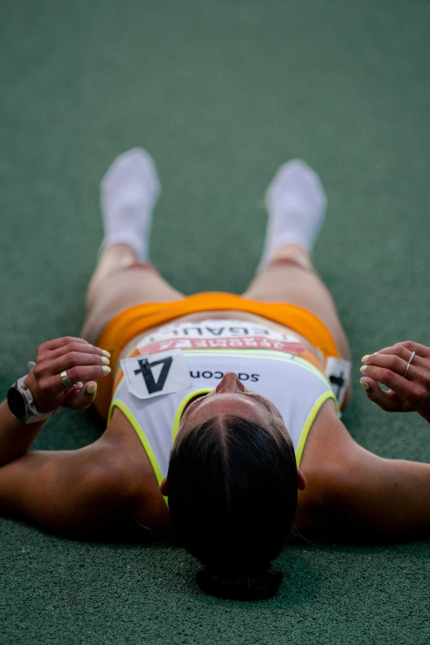 A female athlete lying on a green track after a race, wearing a white and yellow sports uniform with a bib number 4, resting with her arms by her sides.