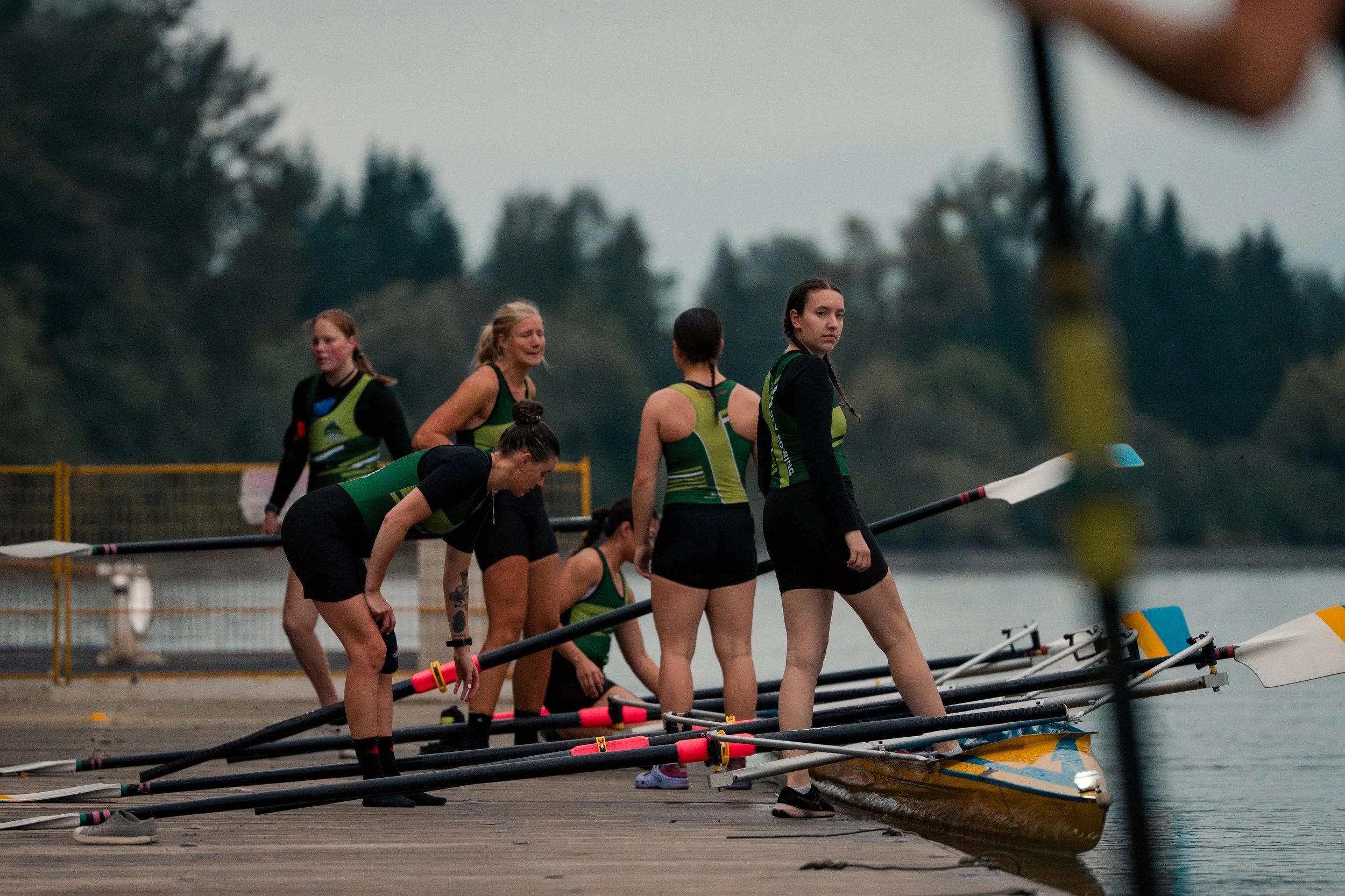 Women in athletic clothing preparing for a rowing race by a calm body of water, some standing, some bending over, with rowing boats and oars on the dock, and trees in the background.