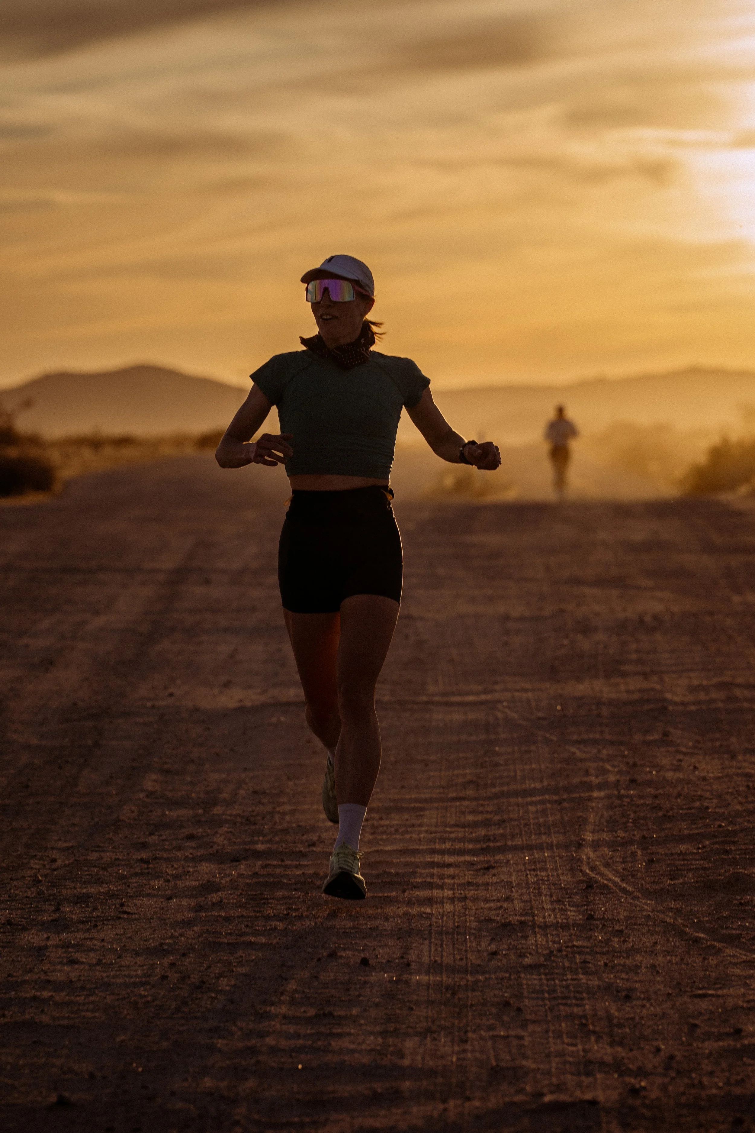 A woman running on a trail at sunset, wearing a cap, sunglasses, and athletic clothing, with a second person running in the background.