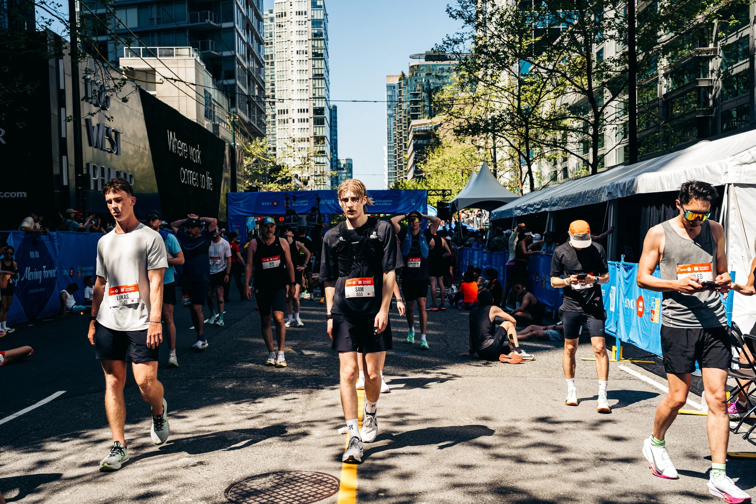 People participating in a marathon race on a city street with tall buildings and blue sky, some walking and others sitting or resting.